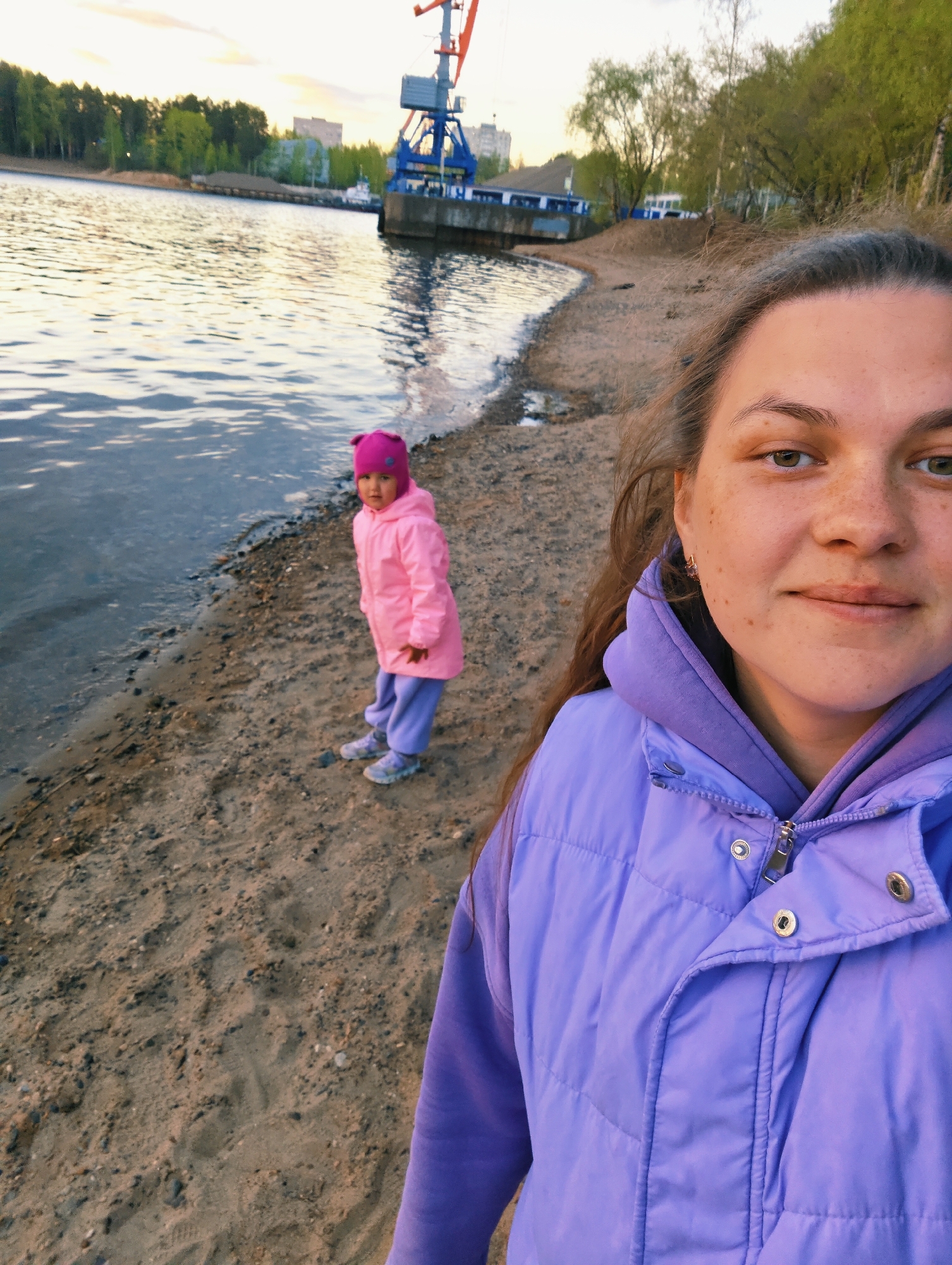 A woman is taking a selfie on a beach in Chaikovsky, Russia. The beach is sandy and next to a body of water. A young girl in a pink jacket and hat stands a few feet away from the woman, near the water. In the background, a blue crane-like structure stands on a concrete platform extending into the water. Trees and buildings are visible on the opposite shore. The sky is light in color, suggesting dusk or dawn. The woman wears a purple puffy jacket over a purple hoodie.
