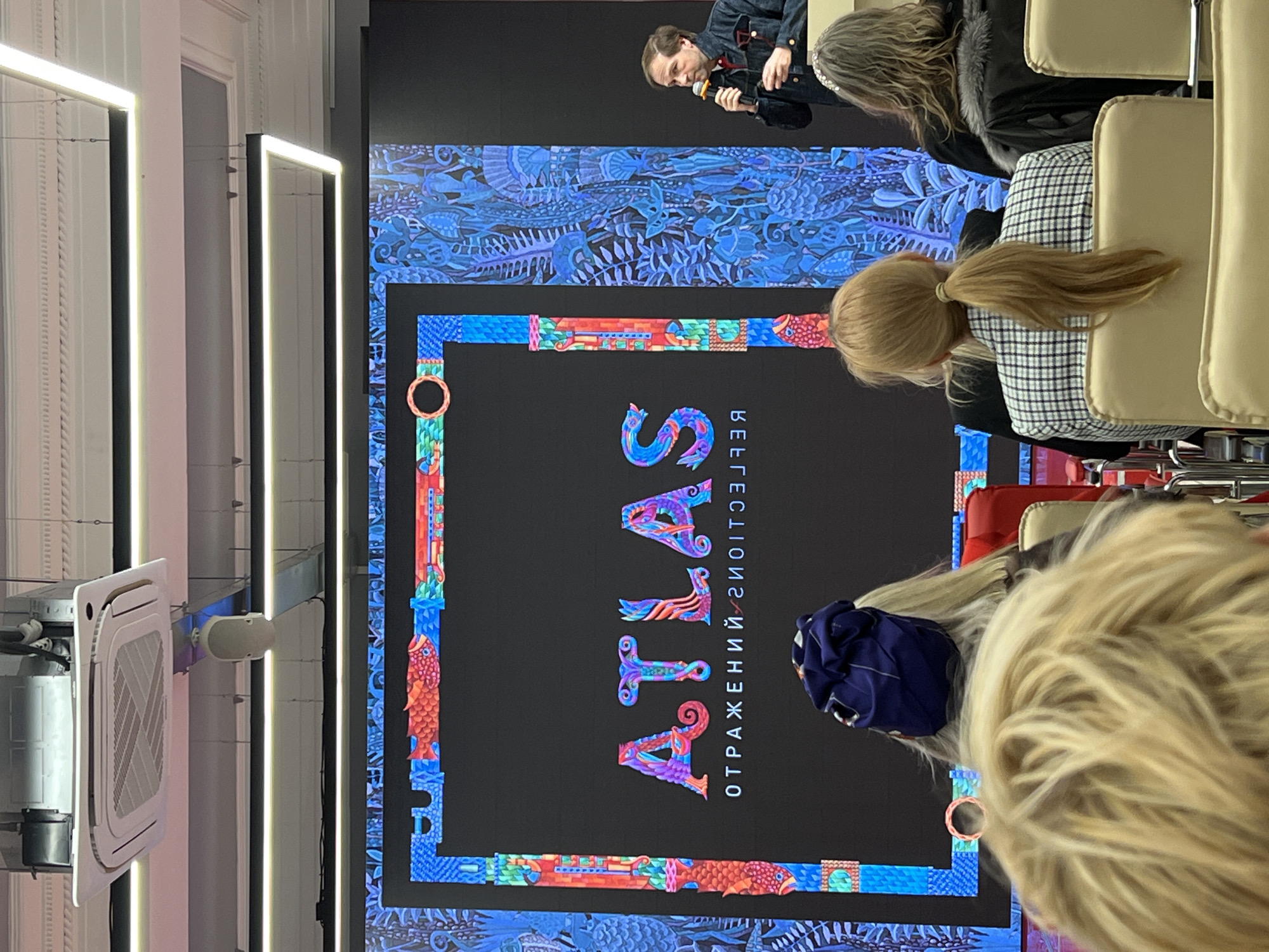 The image shows a presentation in Saint Petersburg, Russia. A presenter speaks into a microphone to an audience. Behind the presenter is a large screen displaying the word "ATLAS" in a stylized font with the word "ОТРАЖЕНИЙ" written in smaller letters. The bottom and top edges of the screen are decorated with patterned imagery. The audience is sitting in chairs. The ceiling has lighting fixtures. Also visible is an air conditioning unit.