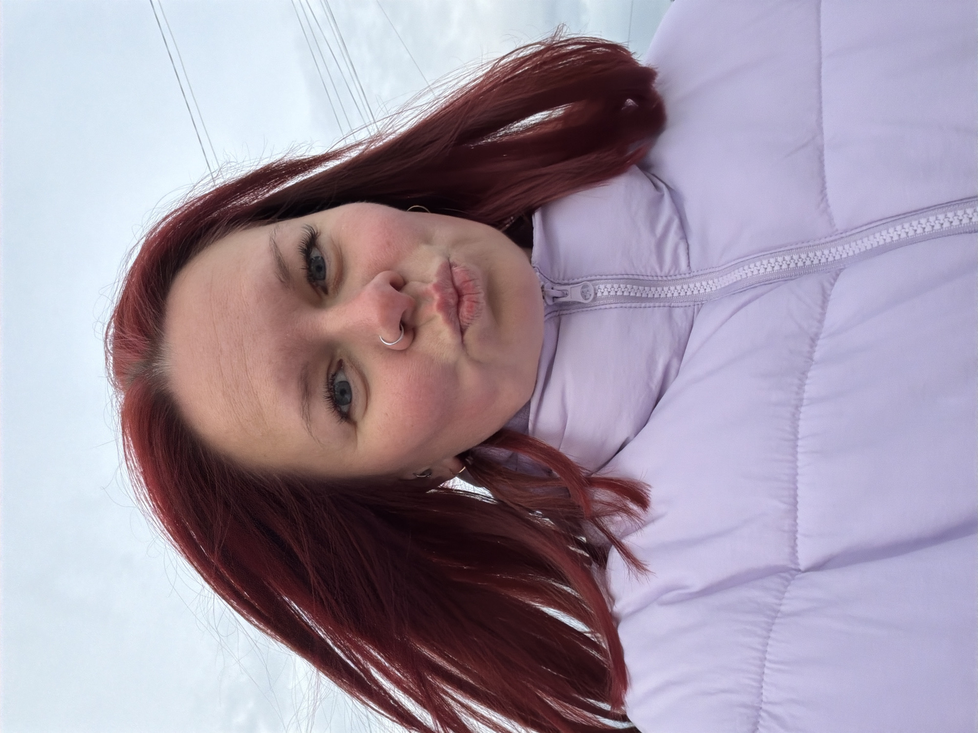 A selfie of a woman with auburn hair and blue eyes, wearing a lavender-colored puffer jacket, with a nose ring, pursing her lips, likely taken outside. Overhead power lines are visible in the sky. The location is indiscernible and cannot be determined to be in the city of Chaikovsky, Russia.