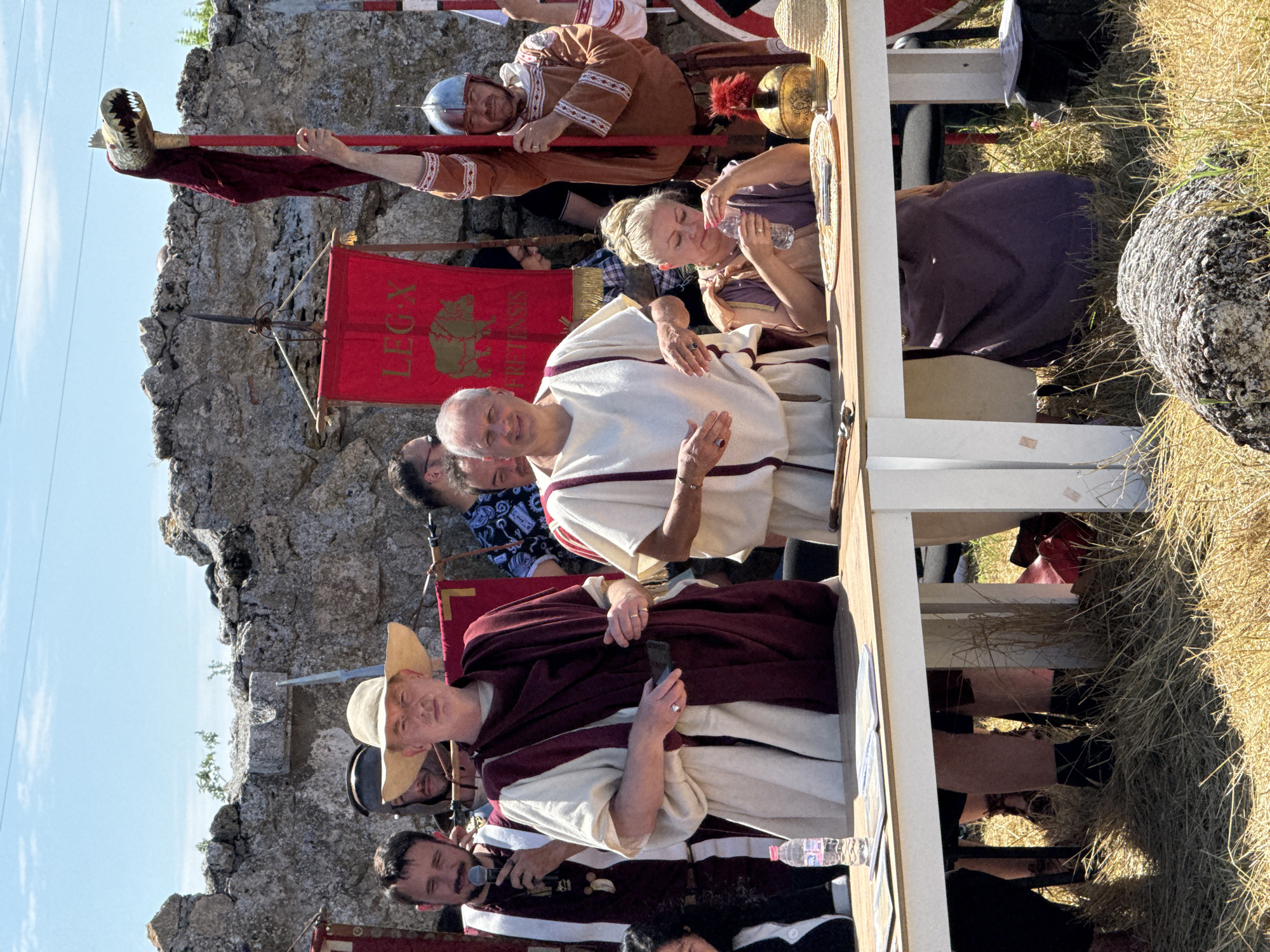 An outdoor scene in Gigen, Bulgaria, depicts a historical reenactment under a clear blue sky. In the foreground, two light-colored wooden tables are positioned on dry, yellowed grass. On the left table, a plastic water bottle and several open documents are visible.
Several individuals are present, many dressed in Roman-style attire. From left to right:
* A man with a beard, wearing a maroon and white toga-like garment, holds a microphone near his mouth.
* Next to him, a man in a wide-brimmed straw hat wears a cream and maroon toga-like garment, holding a dark object in his right hand. A silver ring is visible on his left ring finger.
* To his right, an older man with short grey hair, smiling, also wears a cream and maroon toga-like garment. A ring is on his left ring finger.
* Seated at the right table, a woman with blonde hair tied up wears a purple and tan toga-like garment. She is holding and drinking from a plastic water bottle. Next to her on the table are a golden Roman helmet with a red plume and a straw hat.
* On the far right, a man in a light brown tunic and a Roman helmet holds a tall standard topped with a stylized dragon head.
In the mid-ground, a red vexillum (Roman standard) displaying "LEG X FRETENSIS" and a golden boar emblem hangs from a horizontal bar, supported by poles. The background features a rough stone wall, possibly ancient ruins or a reconstruction. A large, weathered rock is partially visible in the bottom right foreground.