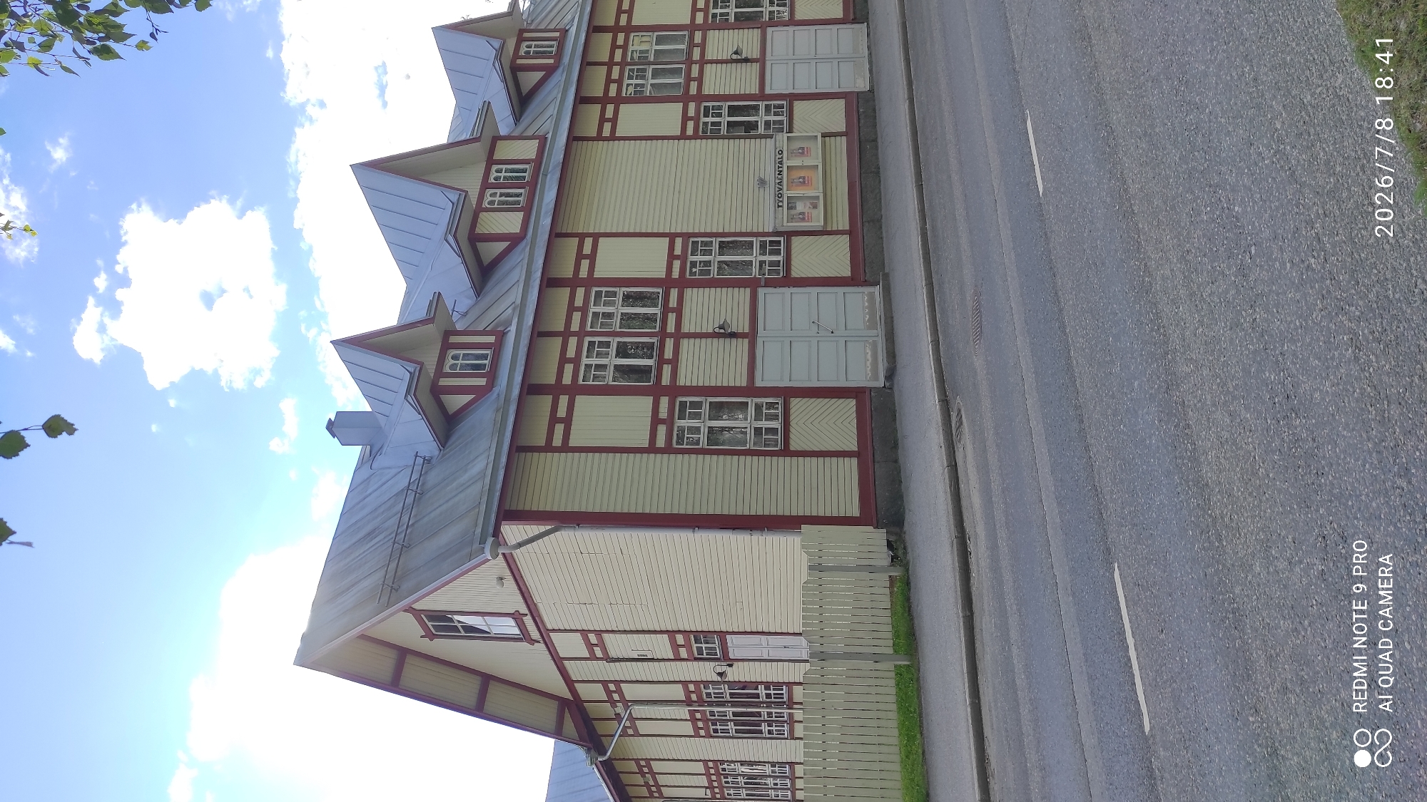 The image shows a two-story wooden building with a metal roof, located on a street in Iisalmi, Finland. The building has a light yellow facade with dark red trim around the windows and along the architectural details. It features multiple windows on both floors, some of which are grouped together. There are several dormer windows on the roof. In front of the building, there is a light-colored wooden fence. A road is visible in the foreground, with markings on the asphalt. There are some posters displayed on the building's facade. The sky is blue with some white clouds, and tree leaves are visible in the upper right corner of the image. The building's signage reads "TYÖVÄENTALO".