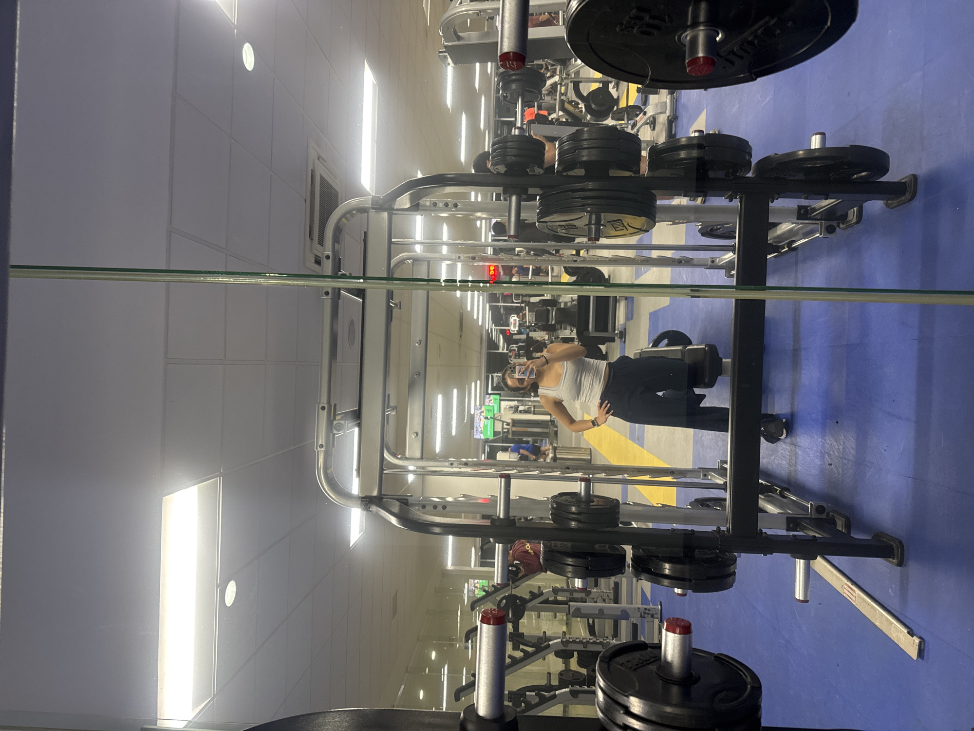 The image shows a mirrored view of a gym setting. The gym has blue and yellow flooring and white walls with recessed lighting. Weightlifting equipment, specifically plate racks and possibly a squat rack, are prominently visible in the foreground. A person, presumably female, is reflected in the mirror, standing and holding a phone. The location in the city of Puebla, Mexico, cannot be confirmed from the image.