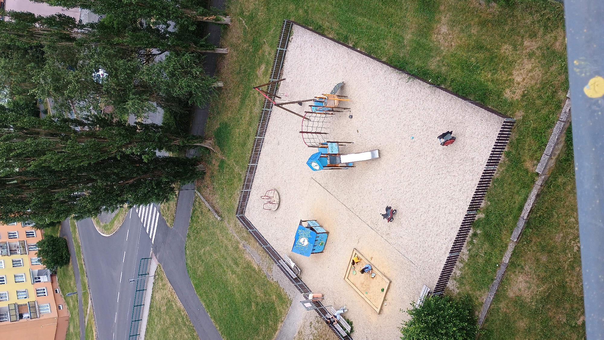 This is a high-angle, medium shot of a modern playground in Sokolov, Czechia. The playground is enclosed by a dark wooden fence and has a sandy surface. In the center of the playground is a blue playhouse with white trim and a red roof. To the right of the playhouse is a multi-colored play structure with a slide and climbing net. To the left of the playhouse is a sandbox with two children playing. There is also a merry-go-round in the background. Two red toy motorcycles are parked near the fence, and a single blue ball is visible on the sand. A bench with three people sitting on it is located outside the fence on the left. The playground is surrounded by green grass and tall trees, and in the background, a modern apartment building with orange and yellow facades is visible. A street with a pedestrian crossing is adjacent to the playground, with a traffic light and a road sign. The scene appears to be on a cloudy day.