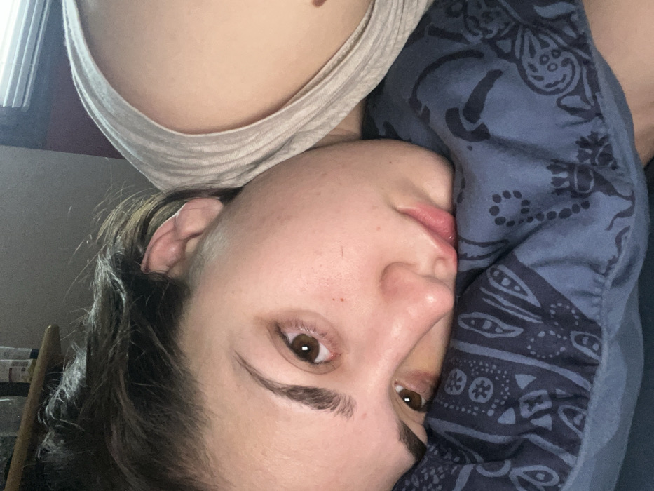 This image captures a young adult female in a reclining position, indoors, located in Ancenis, France. Her head is positioned on a blue pillow displaying a dark paisley pattern. She lies on her right side, with her face oriented towards the camera, her chin resting on the pillow's surface. Her brown eyes are open and maintain a direct gaze. Dark hair is visible along the top of her head. She is clad in a light-colored tank top, which reveals her left shoulder and a portion of her upper chest. A small, dark mole is discernible on her upper chest. The background is out of focus, with a shelf holding containers visible on the upper left, and a window with horizontal blinds visible on the upper right.