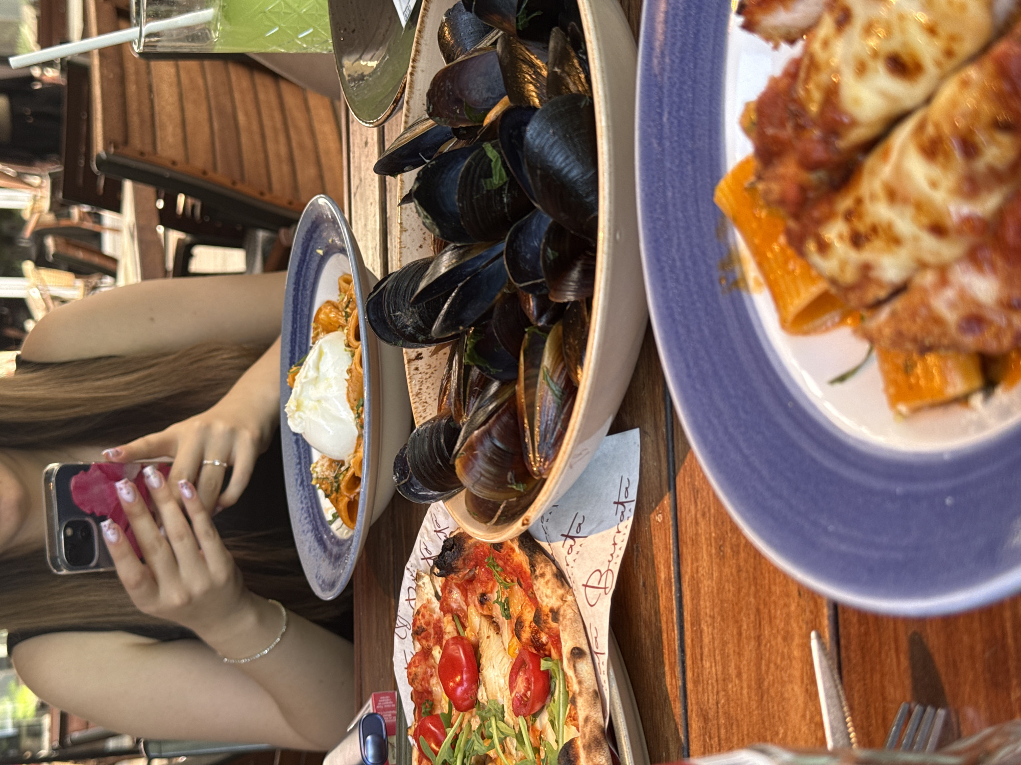 A medium shot captures a table setting at an outdoor restaurant. A person, seen from the torso up, with long brown hair, is holding a smartphone in their hands, engaging with it. Their nails are painted with a light, patterned design. A thin silver bracelet adorns their left wrist, and a ring is visible on their right ring finger. On the table, a pizza with cherry tomatoes and arugula rests on a piece of branded paper. A bowl filled with mussels is positioned next to the pizza. In the background, a plate of pasta with a large dollop of burrata is visible. A glass of green liquid with a straw and another plate with pasta dishes are also present. The table is made of dark wood. The background suggests a busy outdoor dining area with other tables and chairs. The image does not contain any explicit indicators of the city of Sofia, Bulgaria.