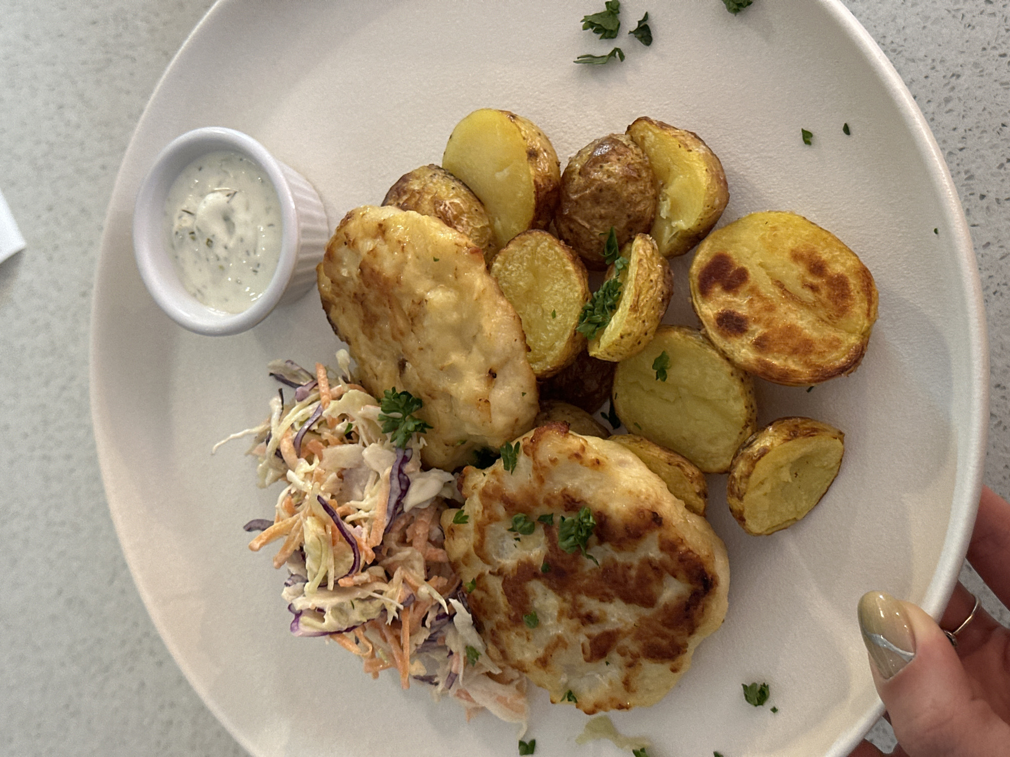 A top-down close-up shows a plate of food. On the plate are two fried patties, a pile of roasted potatoes cut in half, a small portion of coleslaw, and a ramekin of white sauce with herbs. The food is garnished with sprigs of parsley. A hand with painted nails is partially visible on the right edge of the plate. The plate is white and has a wide rim. The background is a light-colored speckled surface. The location is unknown and no information is available to determine if it is in Novi Sad, Serbia.