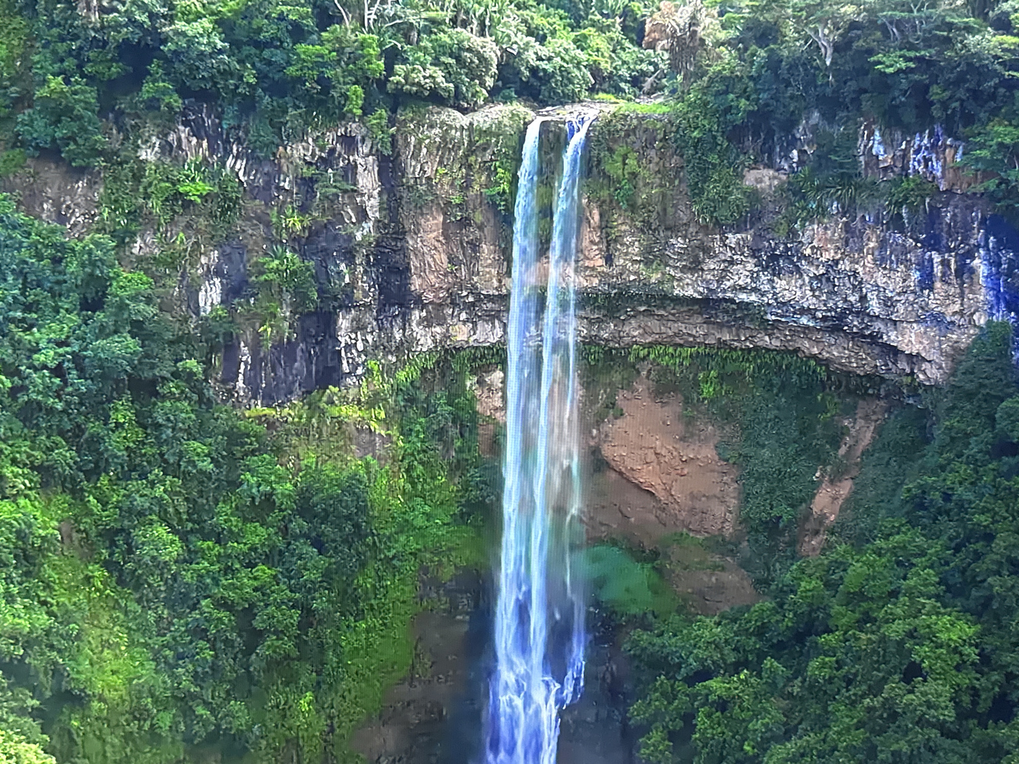 The image shows a tall, multi-tiered waterfall cascading down a rugged, rocky cliff face covered in lush green vegetation. The water appears to be a bright, translucent blue and white as it falls, with mist rising from the base. The surrounding cliffs are a mix of earthy tones and dark rock, with some areas showing streaks of white and blue. Dense green foliage, including trees and smaller plants, frames the waterfall and covers much of the cliffside. The top of the cliff is also adorned with thick greenery, suggesting a natural, remote environment. There are no people or man-made structures visible in the image. The scene depicts a powerful natural phenomenon in a verdant, mountainous landscape. This image does not depict any location in the city of Burgas, Bulgaria.