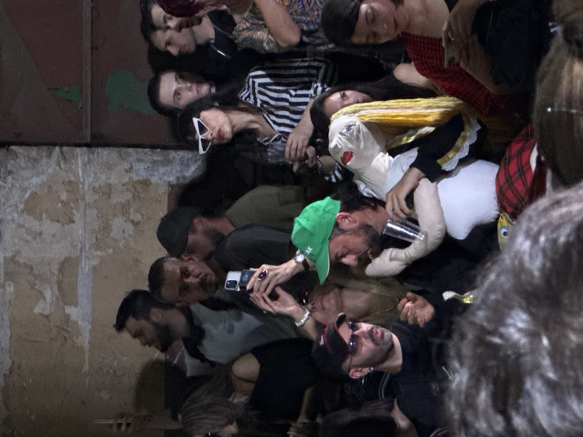 The image depicts a crowd of people in what appears to be an indoor venue, possibly a club or art space. The overall atmosphere is dimly lit with some artificial lighting. In the foreground, several individuals are visible, some in focus and others blurred. One person, wearing a black t-shirt and sunglasses, is looking to their left with an open mouth. To their right, a person in a green cap is holding a clear plastic cup, looking to their right. Between them, a white doll-like figure with red lips and blue eyes is partially visible. Further back, a woman in a striped black and white shirt and angular white sunglasses is holding a drink and a phone. Others are also seen holding phones, suggesting they are documenting the event. The background consists of textured walls and hints of dark structures. The scene is consistent with a social gathering or performance event, potentially in a city like Sofia, Bulgaria, given the context provided, though no specific Sofia landmarks are identifiable within the image itself.