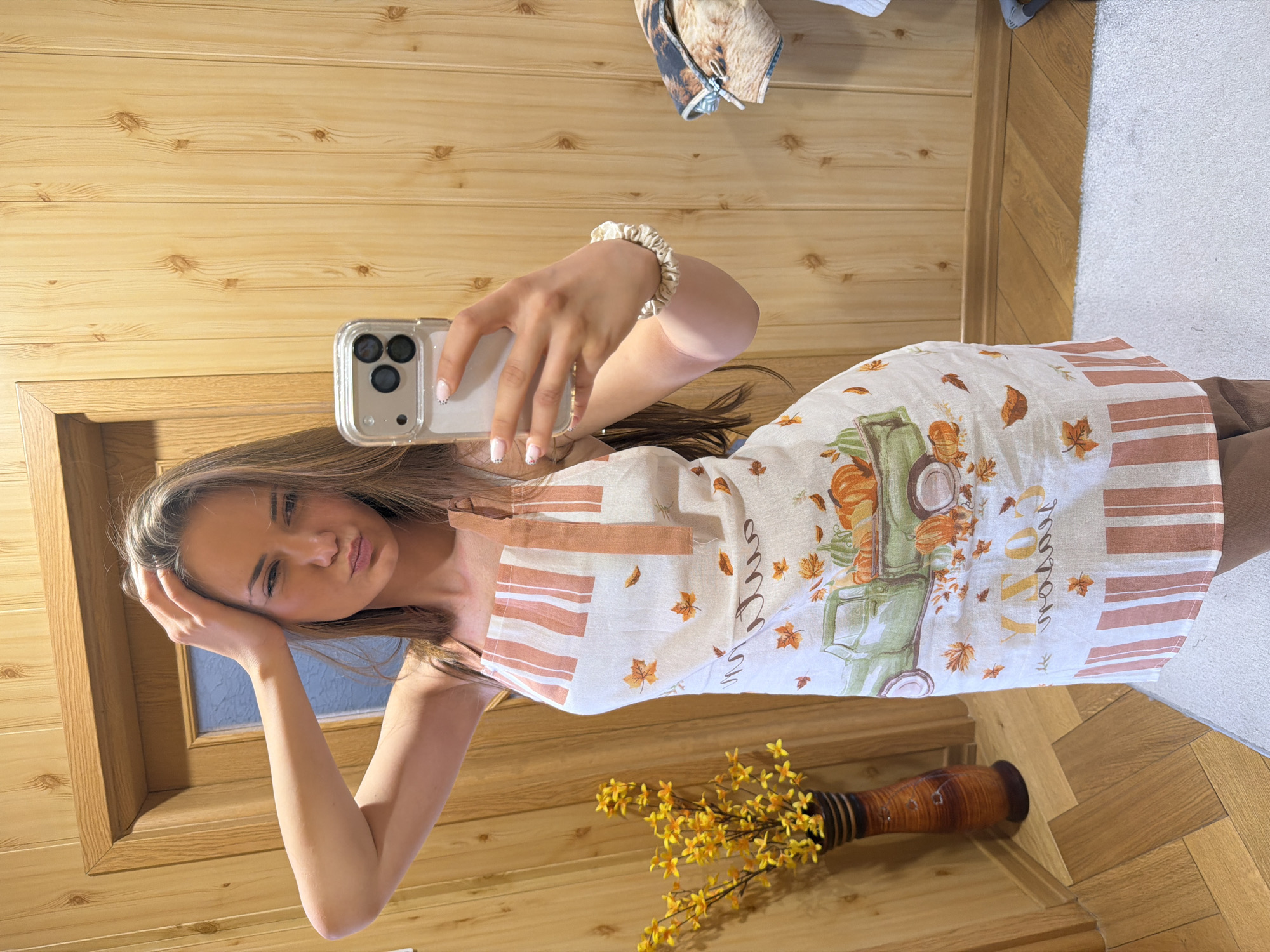A young woman is captured indoors in a selfie, likely in Buhovo, Bulgaria. She has long, light brown hair and a slightly pouting expression as she looks directly at the camera. Her left hand holds a silver smartphone with a clear case and triple camera lenses, while her right hand rests on the back of her head. She wears a light-colored scrunchie on her right wrist, and her nails are painted a light shade with small black dots.
She is dressed in a white or off-white sleeveless garment, resembling an apron or a dress, adorned with an autumnal theme. The design features a vintage-style green pickup truck filled with orange pumpkins and gourds. Scattered brown and orange autumn leaves are printed across the fabric, complemented by rust-colored vertical stripes along the sides. The word "COZY" is visible in decorative script on the garment.
The setting appears to be a brightly lit interior, possibly during the daytime. The background is dominated by light wooden paneling. A wooden door frame is visible to her left. The floor features herringbone-patterned light wood and transitions to a light grey carpet on the far right. A tall, textured brown vase containing bright yellow artificial flowers or branches stands in the lower right corner. A piece of folded fabric with a patterned design is visible in the upper right background.