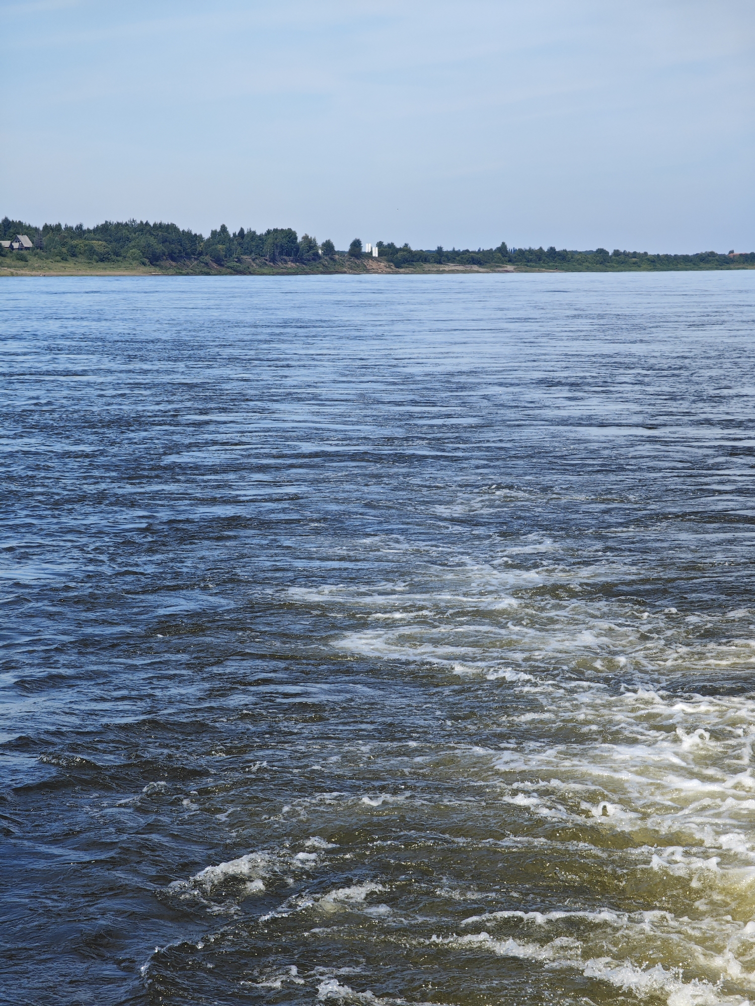 The image captures a wide, calm river under a bright, clear blue sky. The water is a deep blue, with ripples and small waves suggesting movement. In the foreground, the water is disturbed, creating a white foam and wake, indicating that a vessel may have recently passed or is present just out of frame. The opposite bank of the river is lined with a dense green forest. On the bank, there are a few scattered houses, and further in the distance, a tall, slender white structure, possibly a tower or chimney, is visible. The scene is serene and natural, typical of a riverside landscape. The location is the Dnieper River near the city of Yartsevo, Russia, a region known for its wide rivers and forested banks.