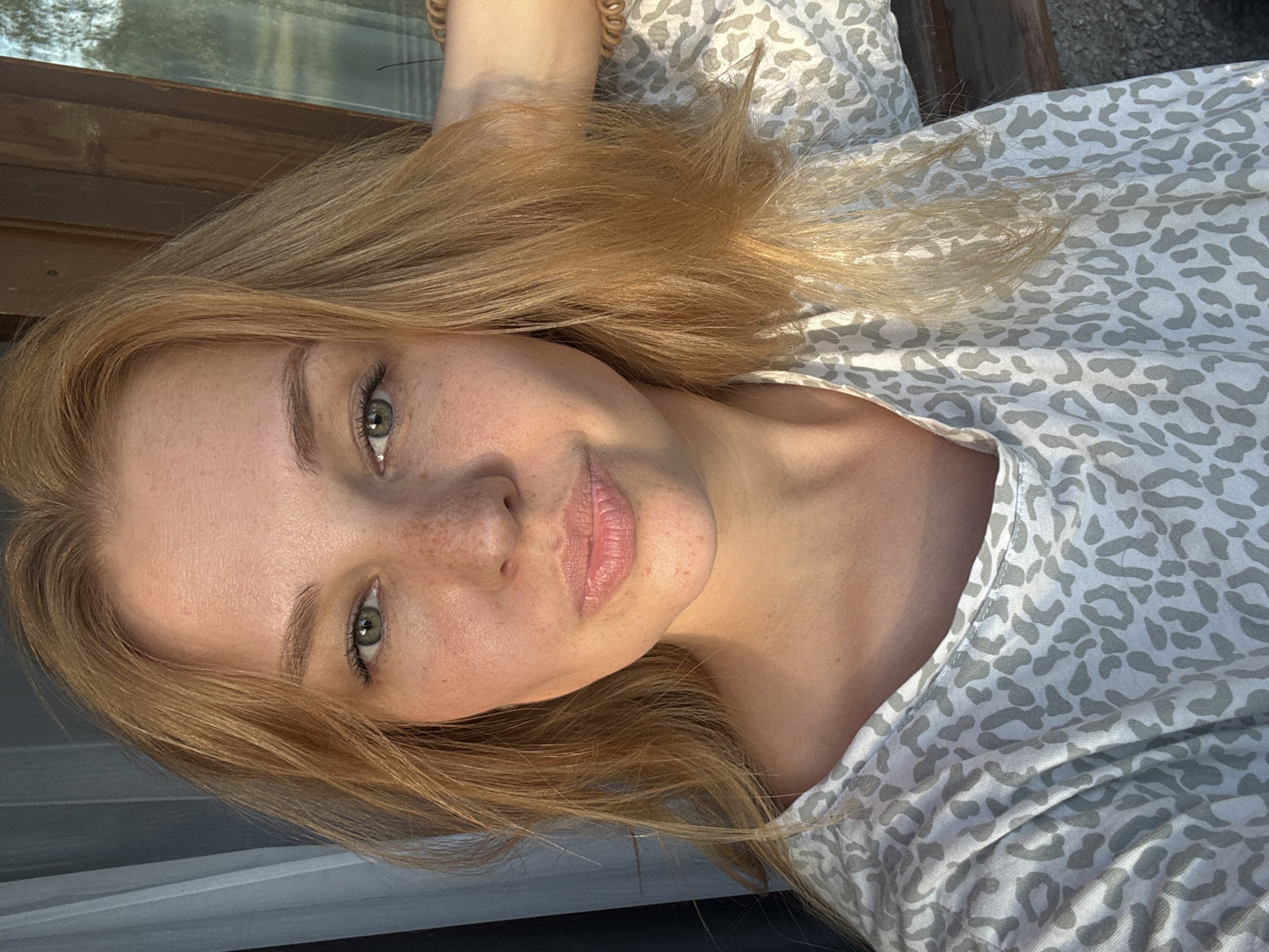A blonde woman with green eyes and freckles is looking into the camera. She is wearing a grey and white leopard print shirt. The background shows a wooden structure and a curtain. The scene appears to be outdoors on a sunny day, possibly in the city of Rusanovo, Russia, given the context.