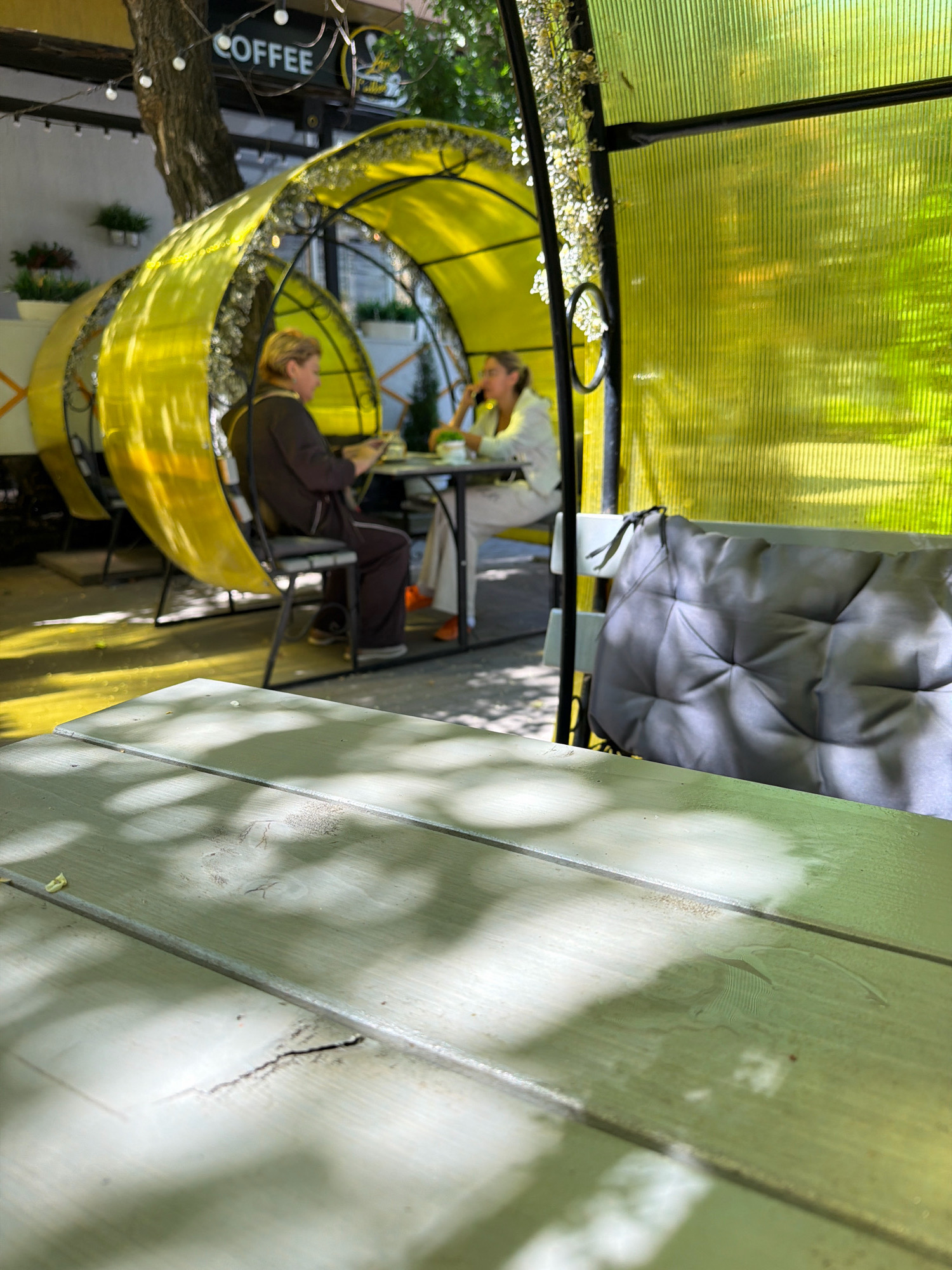 The image depicts an outdoor seating area of a cafe, likely in Yessentuki, Russia, given the context. In the foreground, a weathered, light-colored wooden table is visible with a cracked surface. Sunlight casts dappled shadows across the table. In the mid-ground, two women are seated at a table under a bright yellow, semi-transparent canopy. One woman, on the left, has blonde hair and wears a dark brown jacket and dark pants. She is looking down at a tablet. The other woman, on the right, has dark hair and wears a light-colored jacket and pants. She appears to be talking on her phone. Behind them, a distinctive crescent-shaped, yellow, translucent structure with a metal frame is partially visible, and further back, a sign reading "COFFEE" is mounted. The environment appears to be a sunny day, with green foliage visible in the background and the yellow canopy creating a vibrant, enclosed space. The overall impression is that of a relaxed outdoor dining or cafe setting.