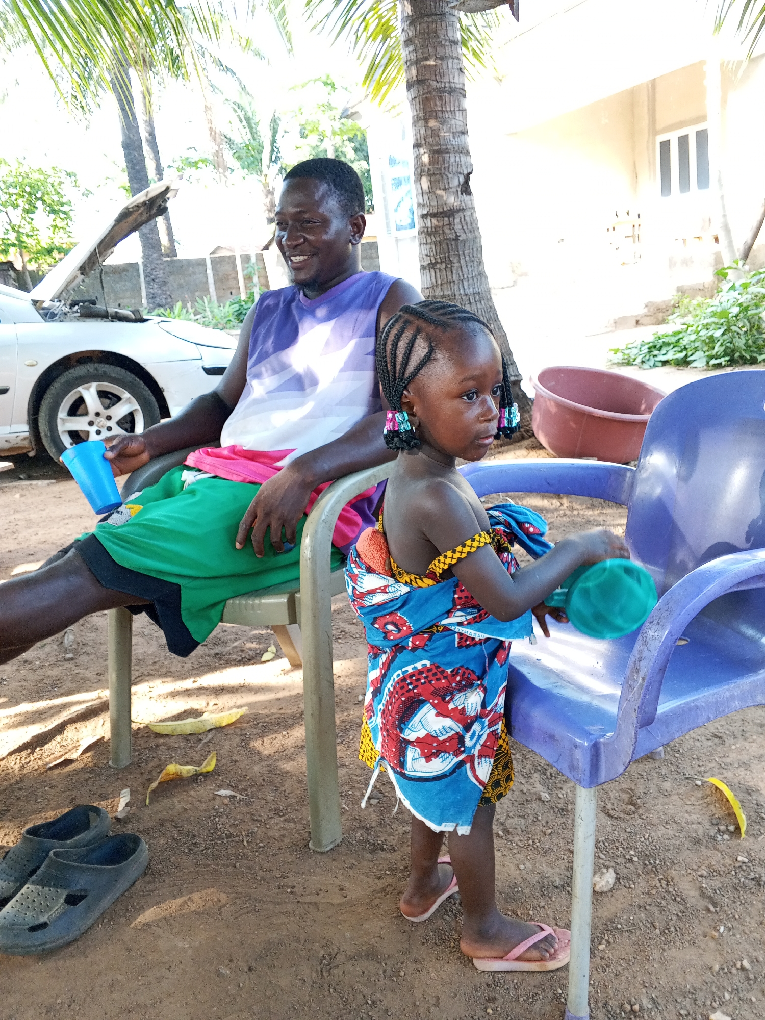 The image depicts a man and a young girl in an outdoor setting, potentially in the city of Unknown, Unknown. The man, dark-skinned, is seated on a chair, wearing a sleeveless shirt and shorts. He is holding a blue cup and appears to be smiling. Next to him, a young, dark-skinned girl is standing, wearing a colorful patterned garment and sandals. She is holding a green object, possibly a toy, and appears to be interacting with a blue plastic chair. In the background, there is a silver car with its hood open, suggesting mechanical work. Additionally, there is a building, some trees, and a maroon-colored basin. The ground is dirt and gravel, with discarded banana peels and a pair of rubber shoes visible near the seated man.