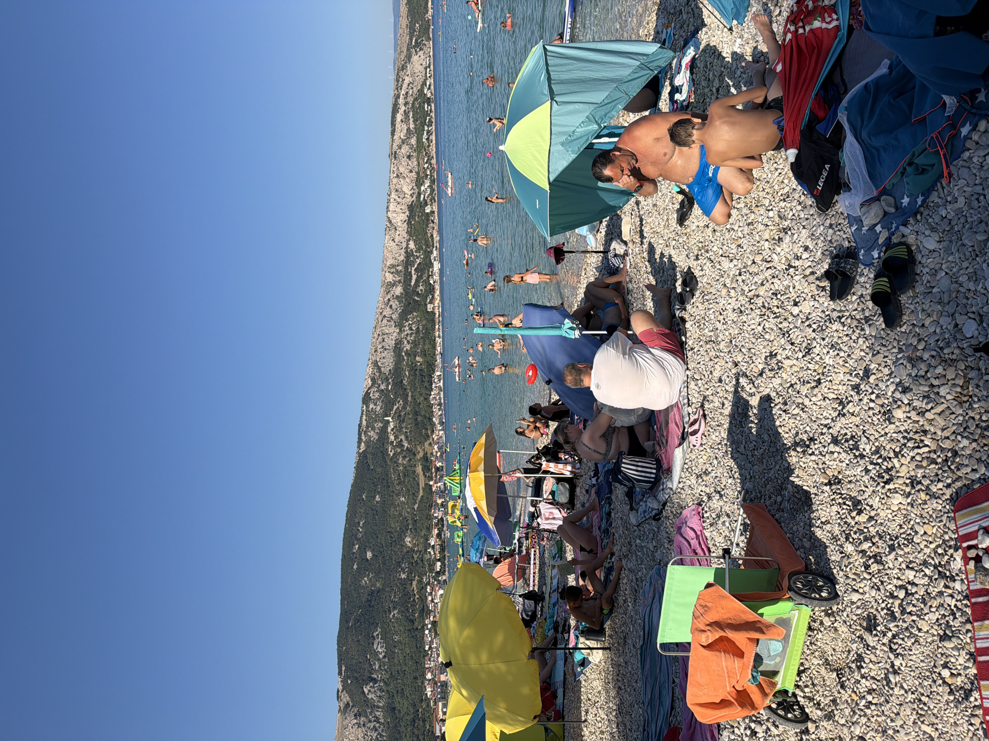 This image depicts a sunny day at a pebble beach in Općina Baška, Croatia. A clear blue sky dominates the upper portion of the frame, with a town nestled at the base of a tree-covered hillside in the background. The sea is calm and filled with people swimming, floating on inflatable toys, and engaging in water activities.
On the beach, numerous individuals are relaxing under colorful umbrellas and beach tents. People are seated or lying on towels and blankets, some reading, others conversing. There are beach chairs and a small wheeled cart with an orange towel draped over it. Footwear, such as flip-flops, are scattered on the pebbles. The foreground shows a concentration of beachgoers enjoying the sunny weather. The scene conveys a typical summer vacation atmosphere.