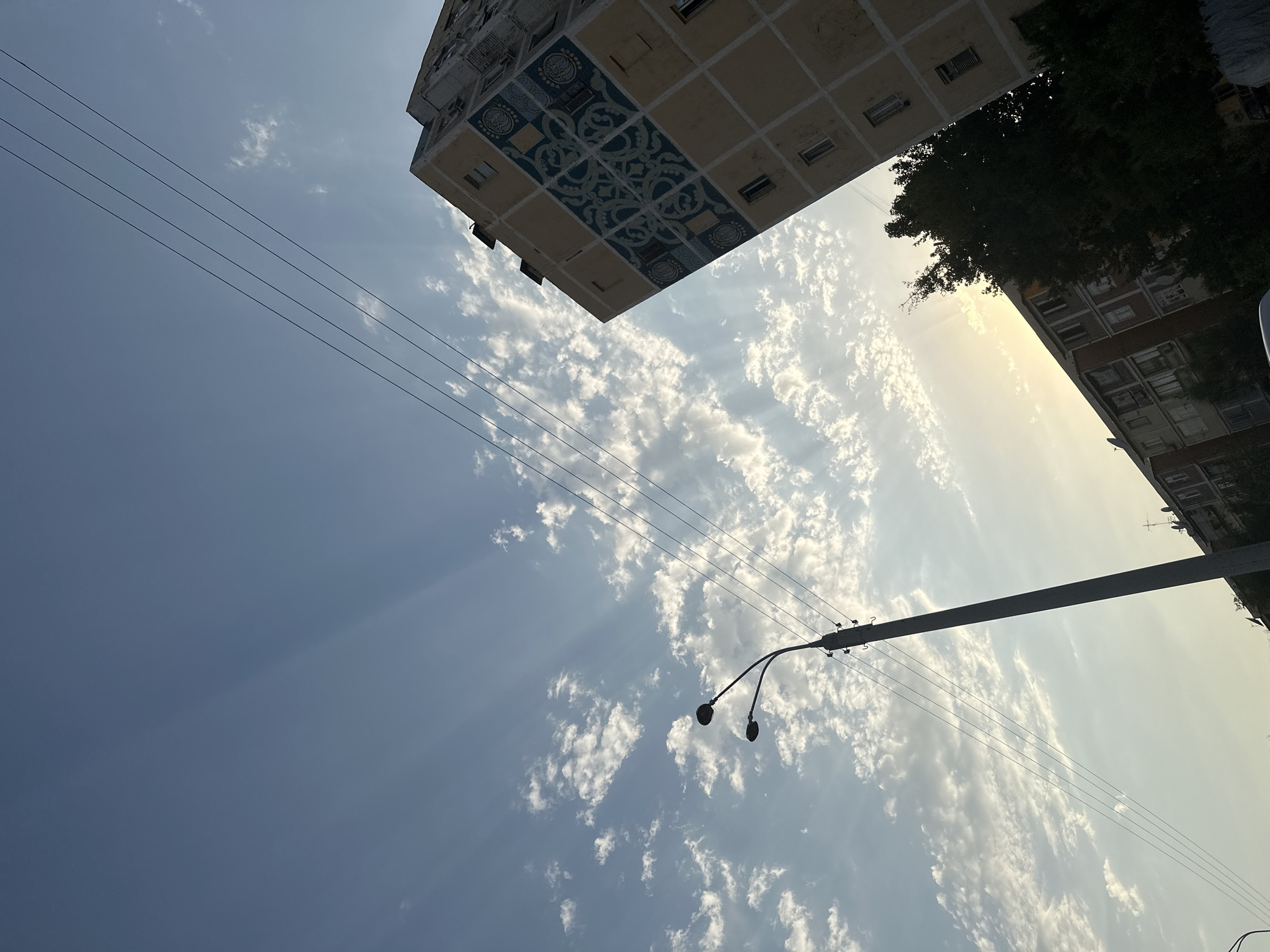 The image captures a view of apartment buildings in Tashkent, Uzbekistan, under a sky streaked with crepuscular rays. A tall streetlamp with two lamps stands prominently on the left, while power lines stretch diagonally across the frame. The facades of the buildings are visible, with one showing a distinct geometric blue tile pattern. Trees and other structures are partially obscured, suggesting an urban environment. The lighting indicates either early morning or late afternoon.