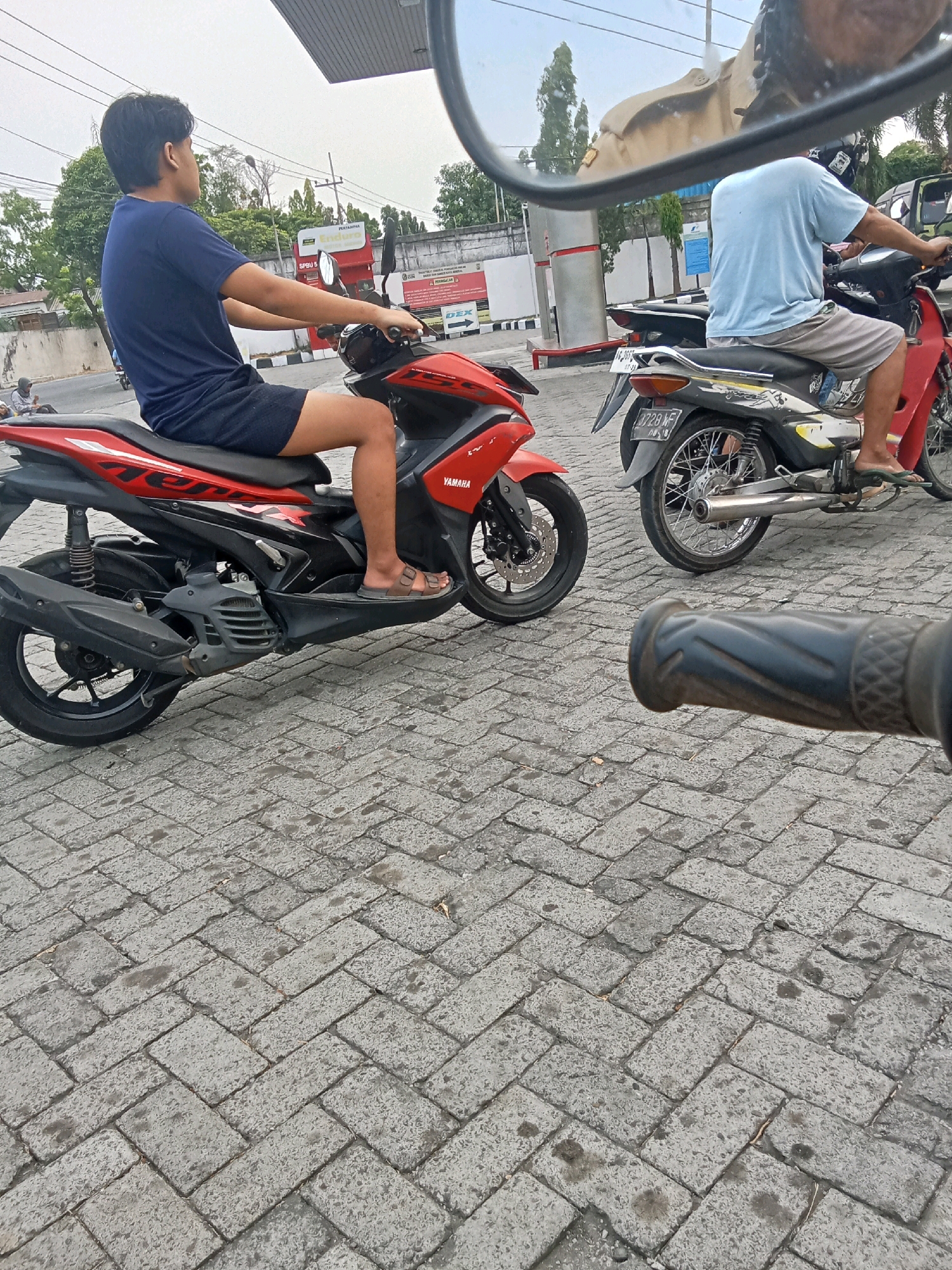 In Jelakombo, Indonesia, two men are pictured on motorcycles at a gas station. The man on the left, wearing a navy blue t-shirt and black shorts, is seated on a red Yamaha scooter. The man on the right, wearing a light blue t-shirt and grey shorts, is on a motorcycle with a license plate that reads "3728 WF". Both men appear to be waiting or observing, with the gas station canopy visible above them and a tiled ground beneath their feet. The scene is captured from a perspective that suggests the viewer is also on a motorcycle, with a handlebar in the foreground.