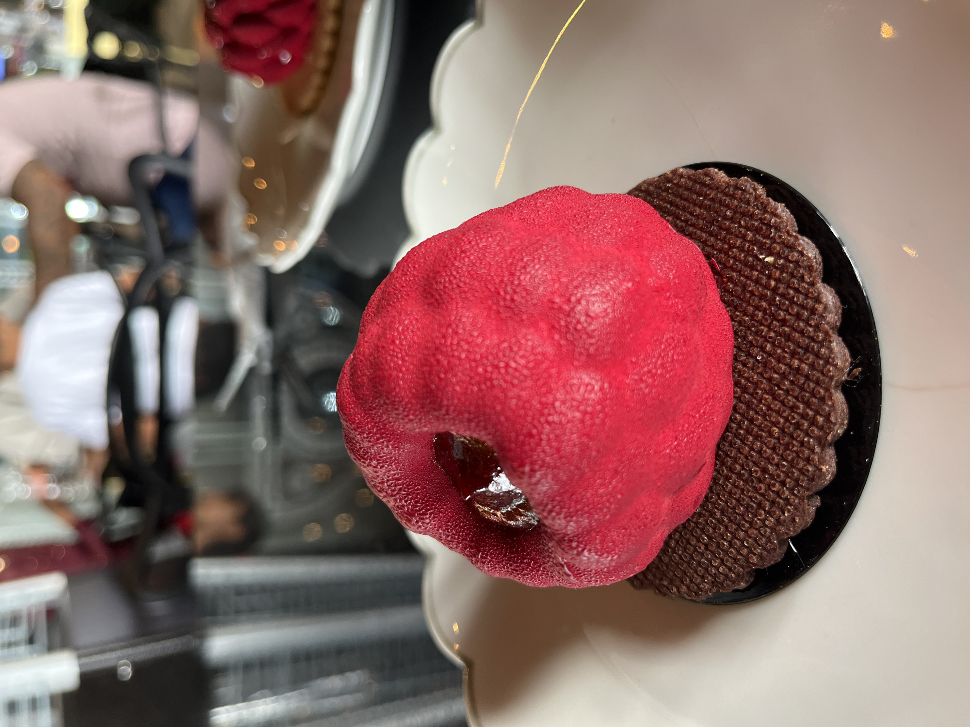 A dessert is presented on a white plate with scalloped edges. The dessert consists of a red, textured dome atop a dark brown, waffle-like base. The plate is placed on a surface with a blurry reflection, likely in a restaurant setting in San Andrés Cholula, Mexico. In the blurred reflection, there are people and objects suggesting an indoor environment.