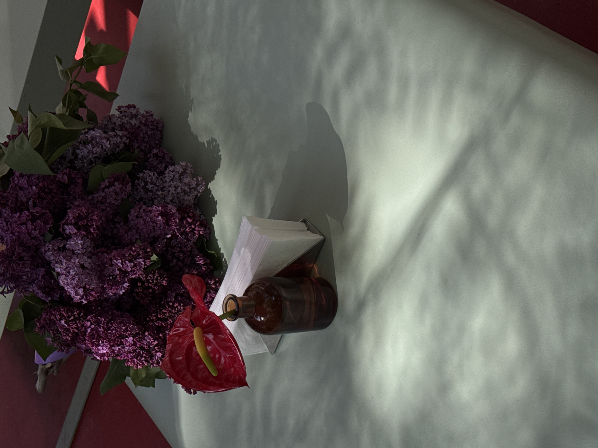 The image shows a table setting in Yerevan, Armenia. On the left side of the table is a bouquet of purple lilacs and a red Anthurium flower. Next to the flowers is a brown glass bottle held in a metal napkin holder containing white napkins. The table surface is a light green color with shadows cast across it. The bottom left corner of the image shows a red surface, likely part of the table.