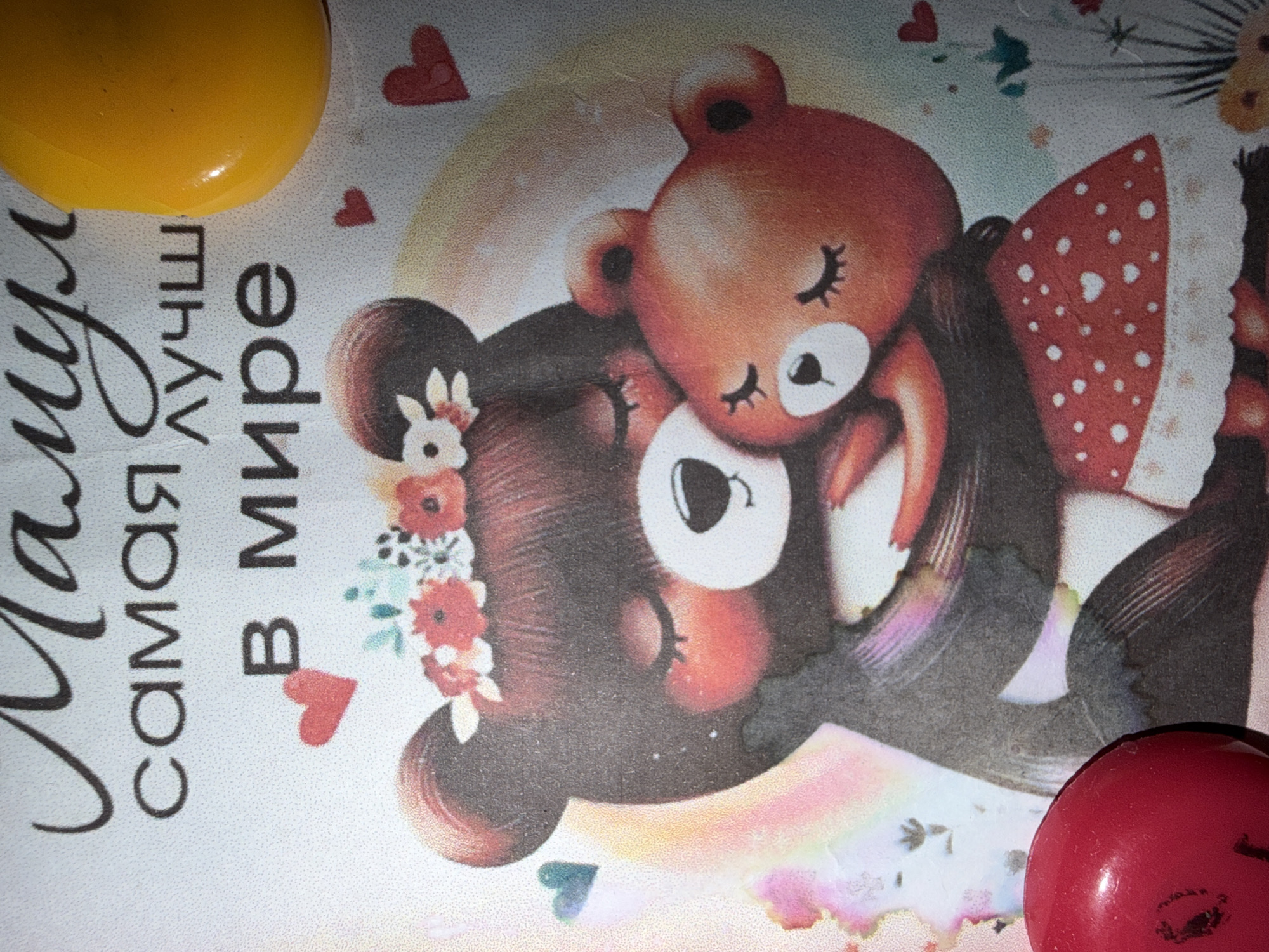 The image shows a close-up of a surface with text in Russian, reading "Маму самая лучшая в мире" (Mom is the best in the world), accompanied by an illustration of two bears. The larger bear, adorned with a floral crown, has a younger bear cub nestled on top of it. The background is a light color with heart-shaped decorations. A yellow object is visible in the upper left corner, and a red object is located in the lower right corner. The location is not determinable from the image.