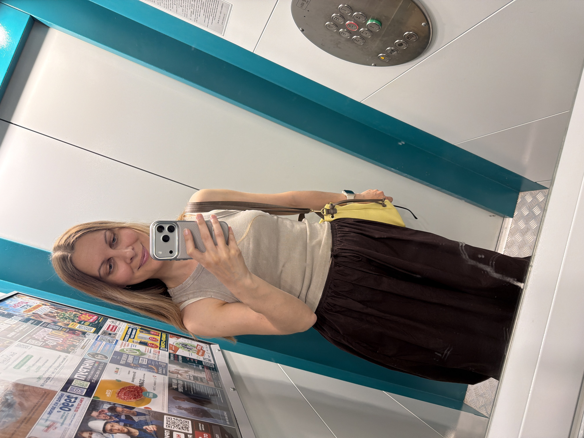 A woman with blonde hair and light-colored eyes takes a selfie in a brightly lit, modern elevator. She is smiling gently at the camera and holding a silver smartphone in her right hand. She is wearing a light-colored, sleeveless top and a long, dark brown skirt. A yellow purse is slung over her left shoulder.
The elevator interior is predominantly white with a teal accent band running horizontally. The floor of the elevator is a metallic diamond plate pattern. On the wall to the woman's left, there is a display of various flyers and posters. Visible text on these flyers is primarily in Russian, with some QR codes present. One flyer shows images of people in uniforms, and another displays food items.
Above the woman's head, in the upper portion of the image, is the elevator control panel. It features a circular metal plate with several round buttons, some illuminated. The buttons have numbers and symbols on them. Text visible on the panel appears to be in Russian.
The overall impression is a candid moment captured by the woman in a public transport setting, likely during the daytime given the bright interior lighting. The mood appears relaxed and casual.