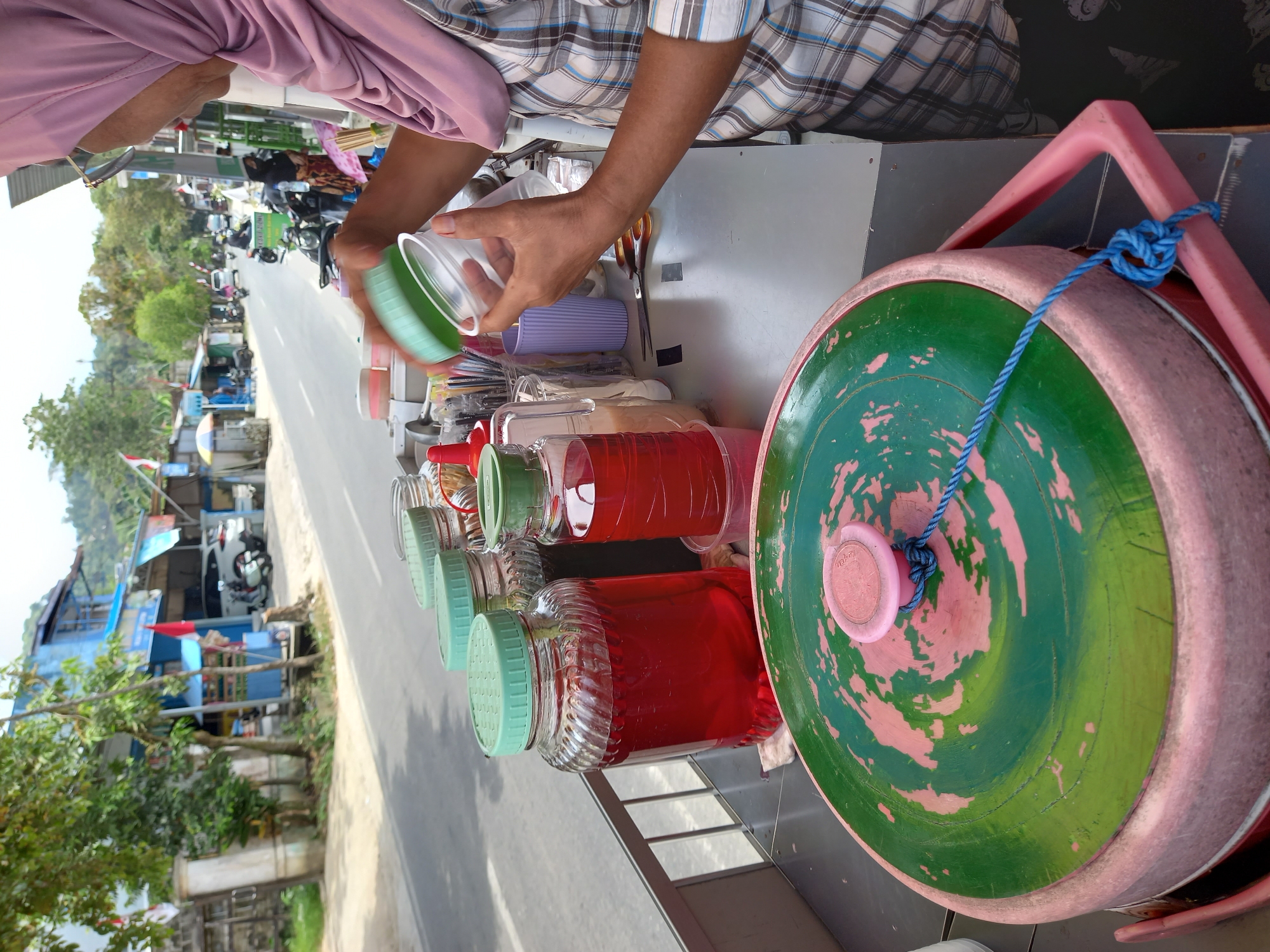 This image depicts a street-side drink stand in Samarinda, Indonesia. A woman wearing a pink hijab and a plaid shirt is in the process of preparing a drink. She is holding a clear plastic cup in her right hand and a green lid in her left, over the cup, suggesting she is about to cover it. The stand features several large glass jars filled with red liquid, likely syrup or a beverage base, and other containers with various ingredients. A large, round cooler with a green and pink lid, secured with a blue rope, is prominent in the foreground. In the background, a road is visible with passing vehicles, including motorcycles and a white car, and buildings lining the street, indicative of an urban or semi-urban setting in Samarinda. The scene appears to be outdoors, under natural light, with a slightly hazy sky.