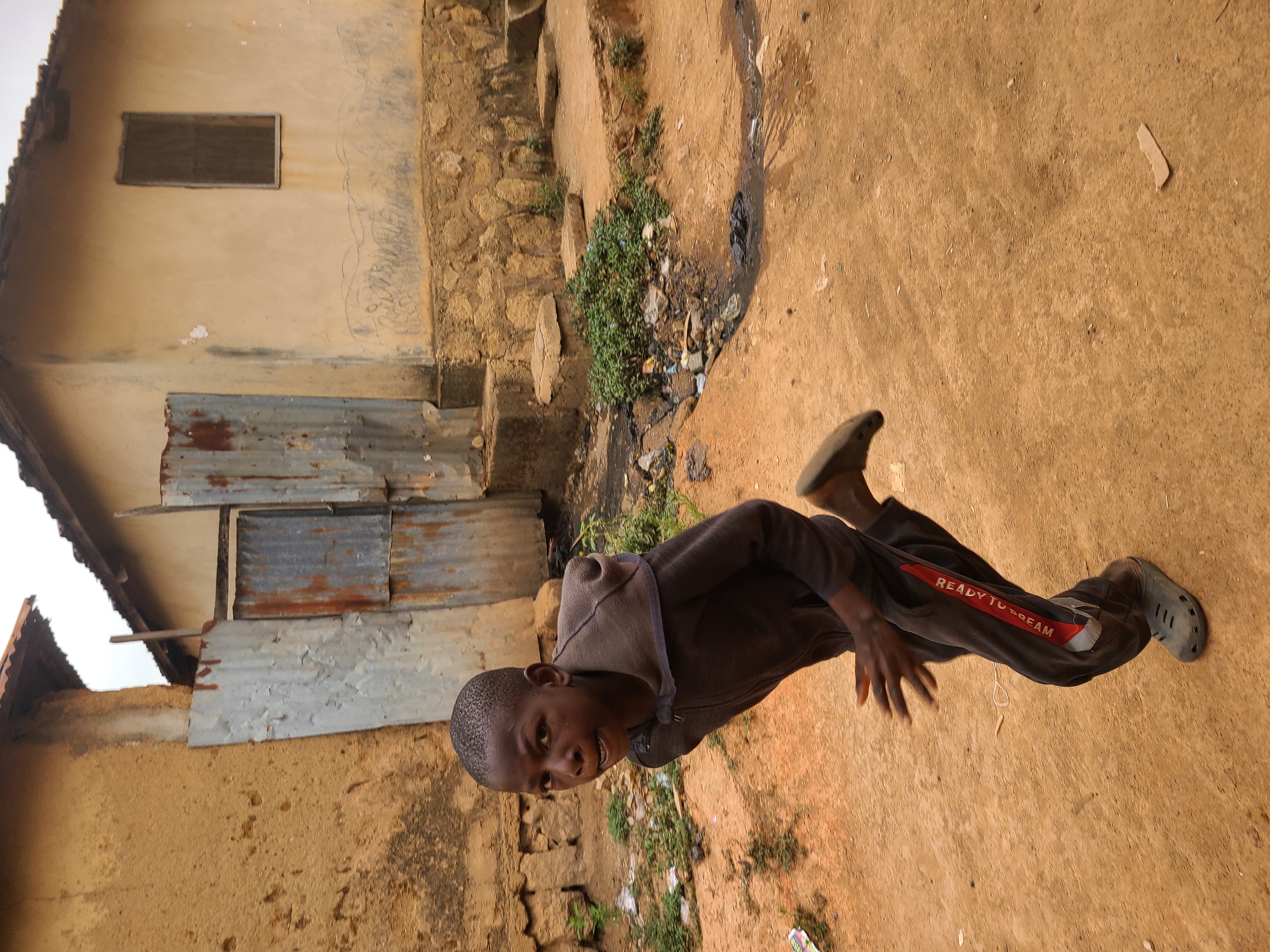 A young boy in a brown hooded jacket and dark pants with "READY TO DREAM" printed on the side, is captured in a residential area in Jos, Nigeria. He is wearing grey rubber clogs. He appears to be standing on a dirt surface adjacent to a building. The building has walls that appear to be made of a light-colored mud or clay, partially covered with pieces of corrugated metal. The roof is partially visible. A small boarded window is visible on the building. The ground surrounding the building is made of packed dirt. There is a drainage ditch with vegetation and some trash. A stone wall also borders the pathway.