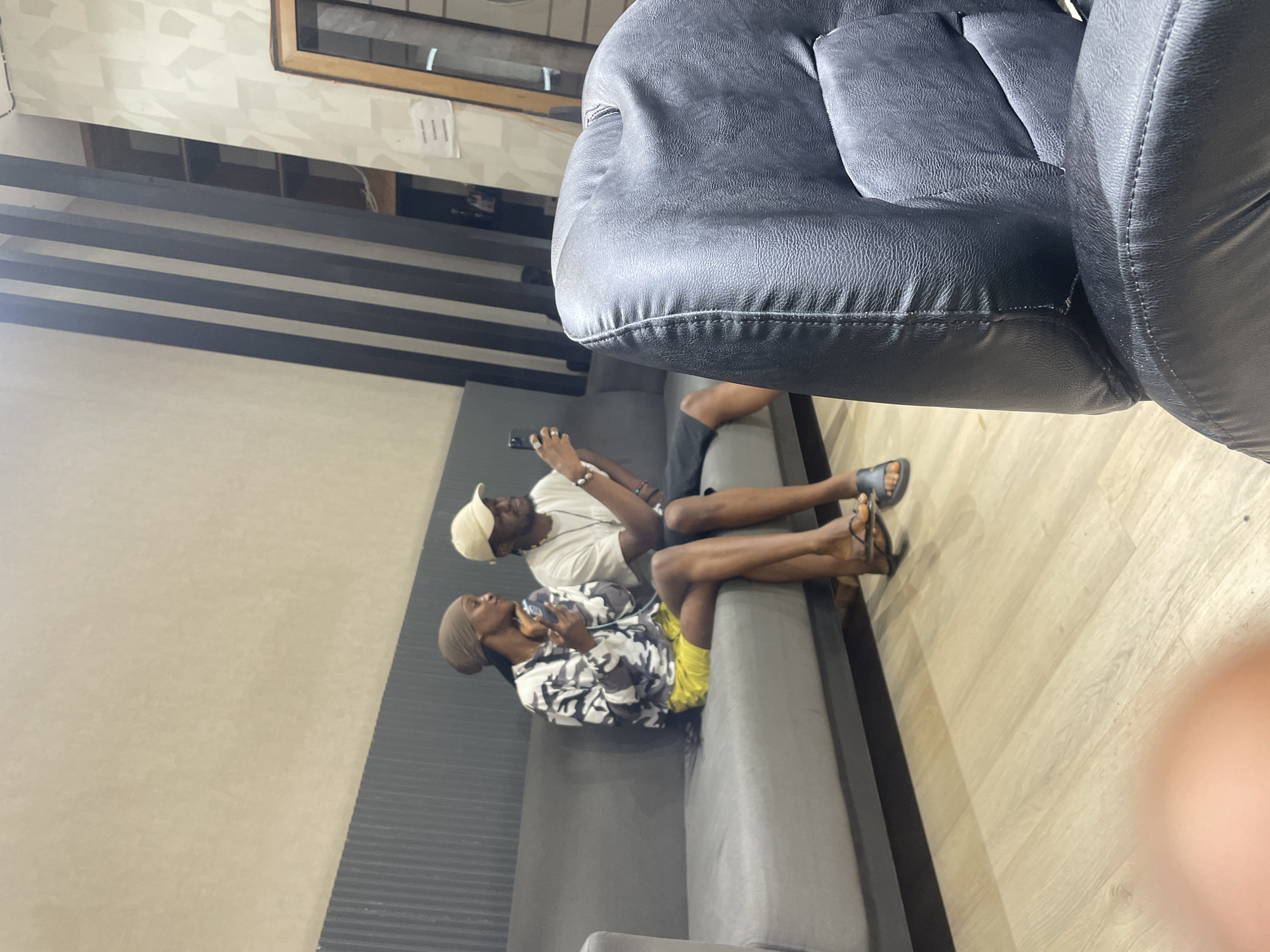 Two individuals, a woman and a man, are seated on a gray sofa in what appears to be an indoor waiting area or lounge. The woman, on the left, wears a headscarf and a camouflage-patterned long-sleeved shirt with bright yellow shorts. She is holding a mobile phone and looking towards it. The man, on the right, is wearing a white t-shirt and a light-colored baseball cap. He is also holding a mobile phone and looking at it. The sofa is placed against a wall with a textured finish and a vertical striped accent. A black leather chair is visible in the foreground, partially obscuring the view of the sofa and the individuals. The flooring is light-colored wood laminate. The environment suggests a contemporary setting, possibly within a commercial or public space in Guzape, Nigeria, characterized by its modern interior design elements.