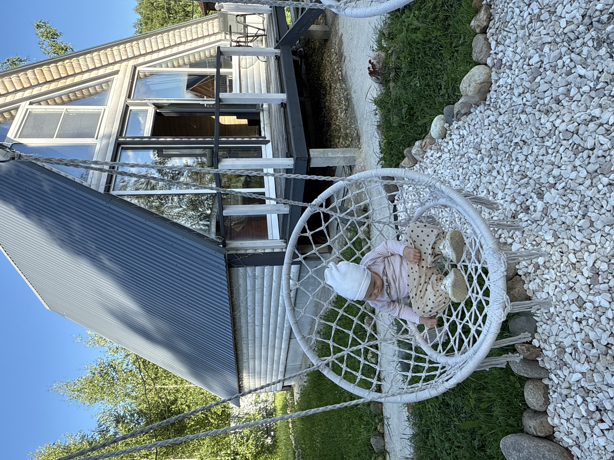 A baby is sitting in a white macrame swing chair in the front yard of a modern A-frame house in Ryzhkovo, Russia. The house is made of wood with a corrugated metal roof and large windows. The baby is wearing a white beanie, a pink and white striped long-sleeve shirt, and beige polka dot pants. The swing is hanging from a rope, and the ground around it is covered in white gravel and grass with some rocks. In the background, there are trees and a clear blue sky.