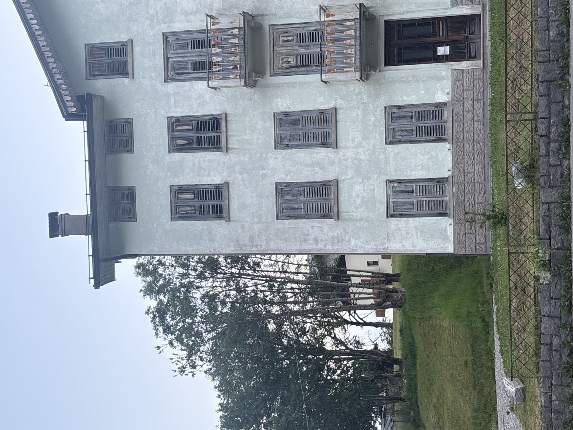 This is a medium-shot, eye-level, exterior photograph of a light blue, weathered building with multiple windows and two balconies. The building has a mansard roof with a chimney and decorative trim. The windows are shuttered with weathered gray wooden louvers. The balconies have white metal railings with decorative orange accents. There is a dark brown double door on the ground floor. To the left of the building, a grassy hill slopes upwards, dotted with trees. A white building is visible behind the trees on the hill. In the foreground, there is a decorative metal fence and a stone wall. The sky is overcast and pale.