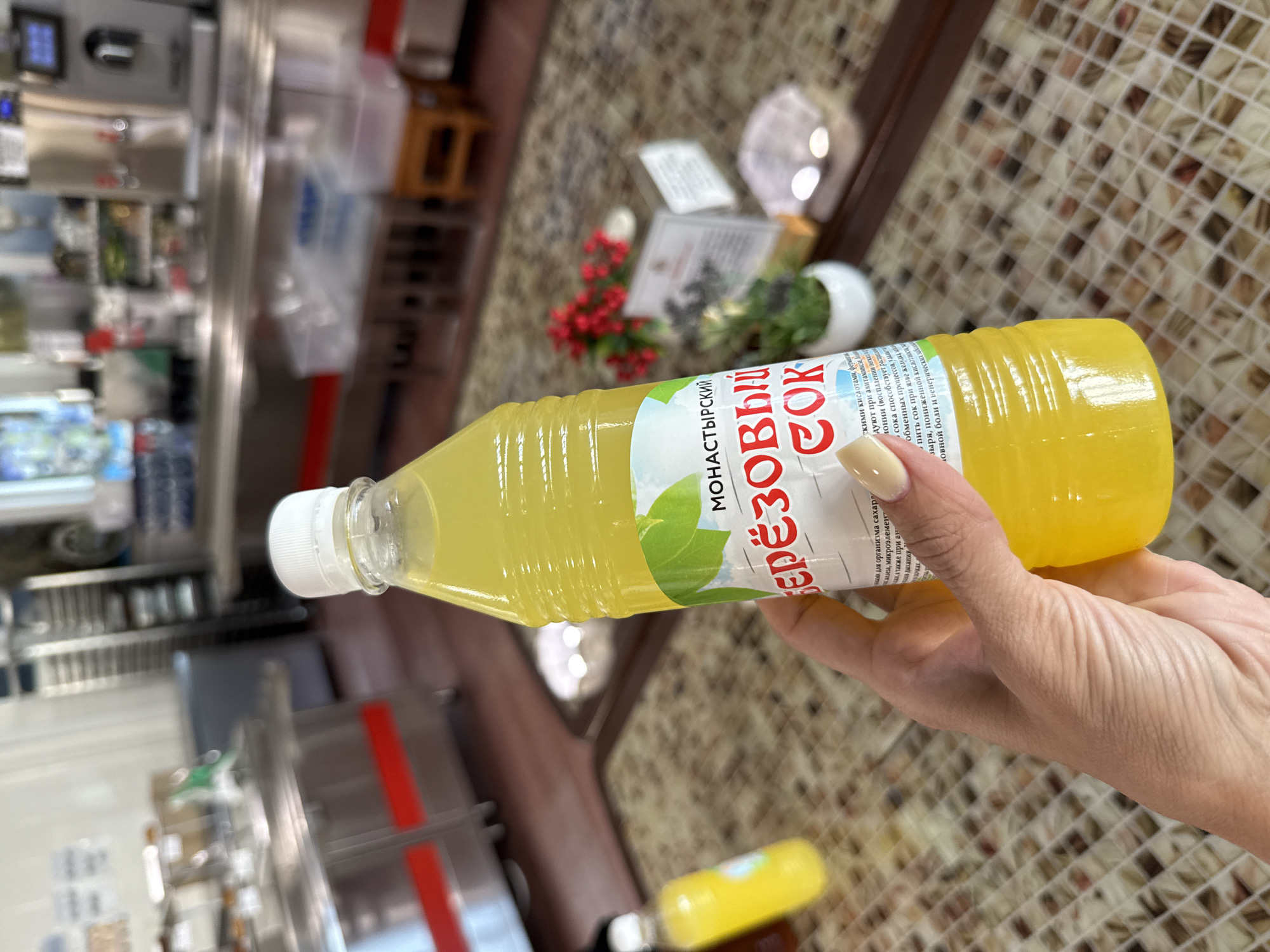 An indoor, close-up, high-angle photograph captures a light-skinned hand with manicured, light beige nails holding a clear plastic bottle filled with translucent yellow liquid. The bottle has a white screw cap and a white label featuring green leaves at the top. The label prominently displays Cyrillic text in red: "БЕРЁЗОВЫЙ СОК" (Birch Sap/Juice), with "МОНАСТЫРСКИЙ" (Monastery) above it, and additional smaller text below. The background is significantly blurred, indicating a shallow depth of field, but reveals an indoor commercial or institutional setting, likely a cafeteria or dining hall. Visible elements in the blurred background include stainless steel serving counters or equipment, some with red accent strips, a wooden stool, another smaller bottle with yellow liquid, small potted plants with red and green foliage, and a white display stand with unreadable text. The floor exhibits a patterned tile design composed of small square tiles in varying shades of brown, beige, and white. The lighting appears bright and even. The location is Unknown, Unknown.