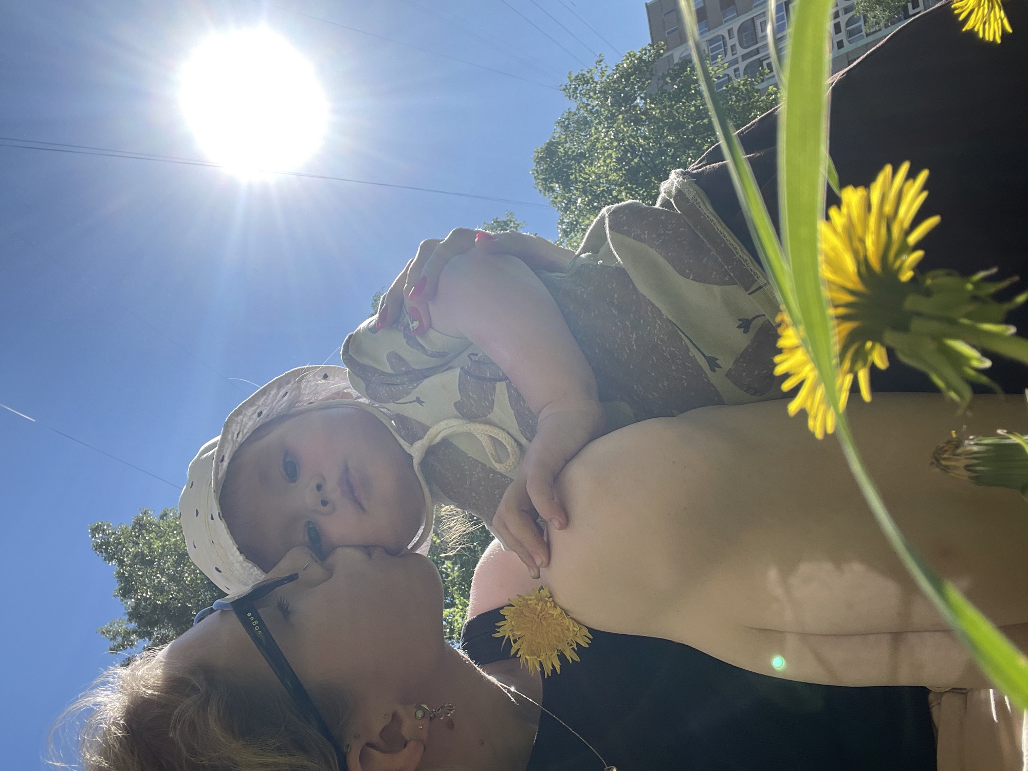 A low-angle shot captures a woman kissing a child on the cheek in Izhevsk, Russia. The sun is brightly visible at the top, casting a glare. The child wears a patterned outfit and a hat. The woman wears sunglasses and has multiple earrings. In the foreground, yellow dandelions are visible. In the background, trees and a building are present.