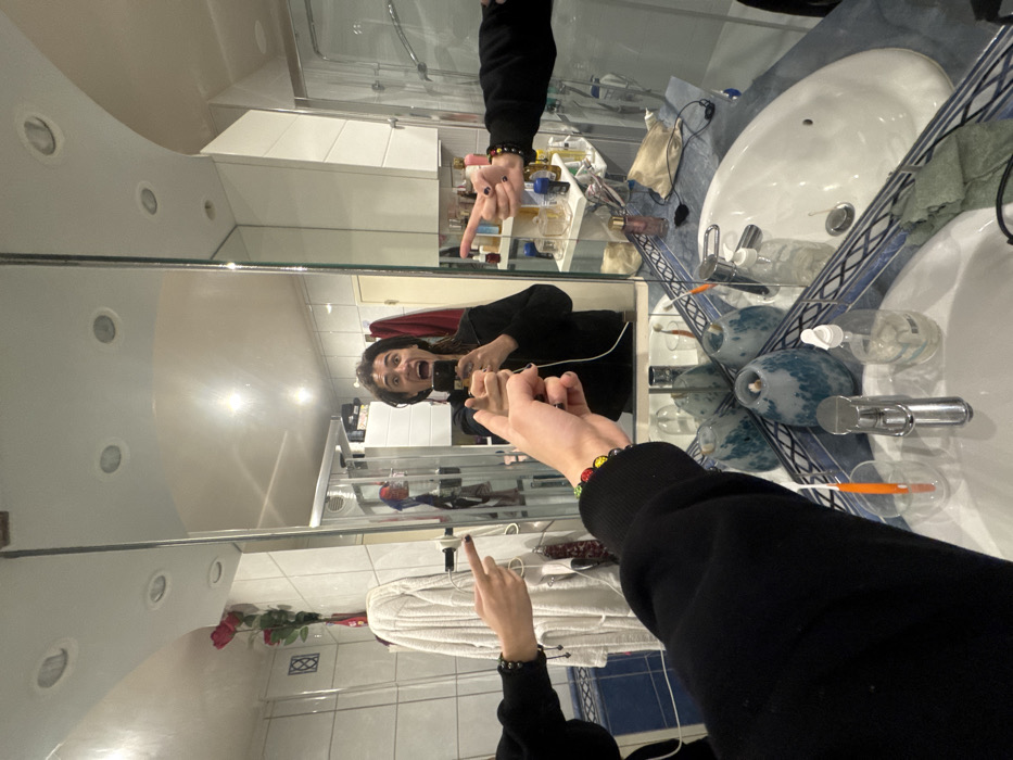 The image depicts a mirror selfie taken in a bathroom, rotated 90 degrees clockwise from its likely original portrait orientation.
In the center of the frame, reflected in a large wall-mounted mirror, is a person with dark hair, wearing a black long-sleeved top. Their face is captured in an expressive, candid moment, with their mouth wide open as if mid-shout or scream, and eyes wide. They are holding a smartphone in their left hand, which is visible in the reflection, presumably taking the picture. Their right hand is extended forward, with the index finger pointing directly towards the camera, while the other fingers are curled into a fist. On their left wrist, a colorful beaded bracelet with red, yellow, green, and black beads is visible. On their right wrist, a simpler, dark-colored bracelet (possibly made of cord) is worn.
In the foreground, the actual left hand of the person (the one holding the camera) is visible, also extending towards the mirror, with its index finger pointing. This creates a visual interplay between the real hand and its reflection.
The background, also seen through the mirror's reflection, is a bathroom setting. To the upper right, a white ceramic sink with a metal faucet is visible, set against a wall with blue and white patterned tiles. The countertop surrounding the sink holds various bathroom items, including bottles of shampoo or soap, a pump dispenser, a toothbrush in a holder, and small containers or perfume bottles. A beige cloth is also present on the counter. The ceiling above the mirror is white and features multiple recessed circular lights. White tiled walls are visible in the reflection, along with a glass shower enclosure or partition. Further down in the reflection, a white folded towel and a single red rose or flower are faintly visible. The overall lighting appears to be artificial, likely from the bathroom's fixtures.
The activity captured is a playful, expressive mirror selfie in a domestic bathroom setting. No discernible text is visible in the image.