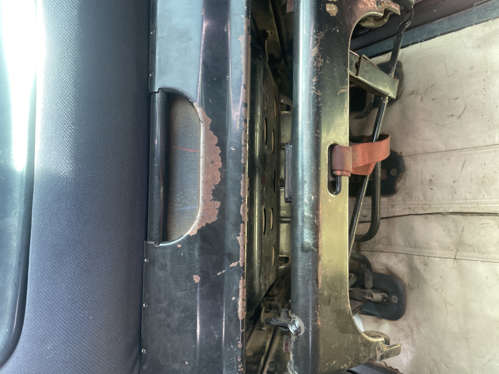 The image shows a close-up, interior view of a vehicle's seating area, likely in Bauchi, Nigeria. The primary subjects are the fabric seat and the metal frame of the seat mechanism. The seat itself is upholstered in a dark, textured fabric. The metal components of the seat are dark, with areas of rust and peeling paint visible, suggesting age and wear. A red fabric strap is also visible, likely part of a seatbelt or securing mechanism. The background shows a white, textured surface, possibly the interior wall of the vehicle, with some grime and marks.
There is no discernible activity or event taking place; the image appears to be a static shot of the seating structure. Notable details include the significant rust and wear on the metal frame, the texture of the seat fabric, and the presence of the red strap. The lighting suggests it is daytime, with a bright light source casting reflections on the metal and fabric. The location cues (Bauchi, Nigeria) are provided but not visually evident in the image itself, other than perhaps a general sense of the type of vehicle or its condition, which could be common in various regions. No visible text is present in the image.