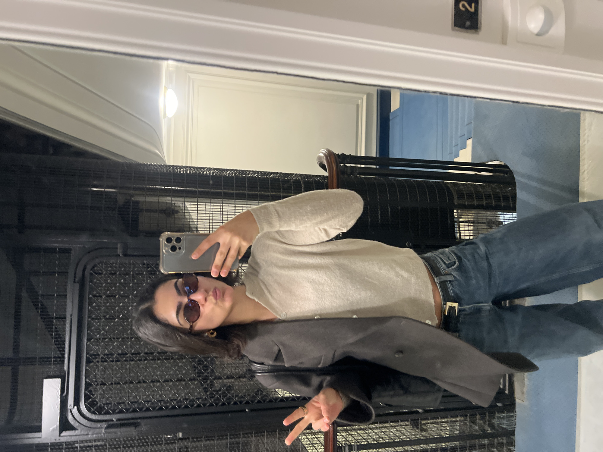A mirror selfie shows a young woman inside an elevator, likely in Paris, France, as indicated by the location context. She is holding a silver-colored smartphone with multiple cameras, possibly an iPhone, to capture her reflection. The woman wears dark, rounded sunglasses, has dark hair, and is pouting her lips. Her left hand forms a "peace" sign, adorned with rings, while her right hand holds the phone.
She is dressed in a light, possibly beige or off-white, long-sleeved cardigan with buttons, paired with blue denim jeans secured by a dark belt with a gold buckle. A dark, possibly black or charcoal gray, jacket is draped over her left arm. A gold-colored hoop earring is visible on her right ear.
The elevator interior features dark metal grates or mesh on the walls and door, creating a textured pattern. In the reflection beyond the woman and elevator structure, a classic architectural setting is visible. This includes white walls with ornate molding, a round white light fixture emitting a bright glow, and a prominent staircase with blue risers and white treads. A small black plaque with the white number "2" is visible near the top right of the reflection, likely indicating a floor number. The overall impression is one of an elegant, possibly older building.