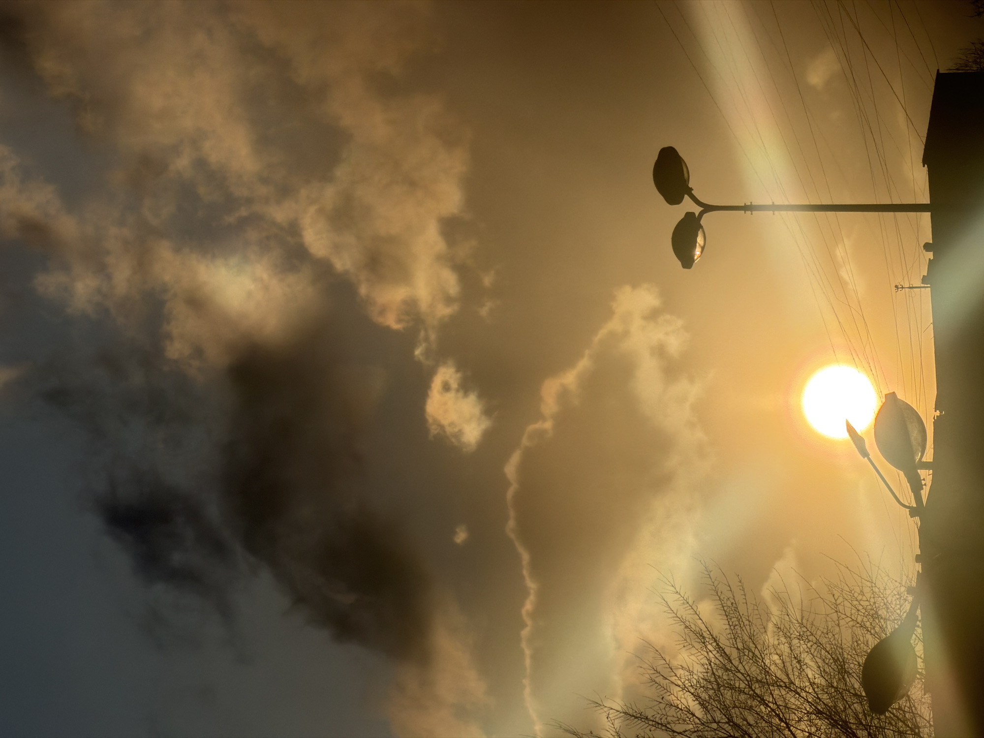 The image shows a low-angle view of the sky, dominated by thick, dark clouds illuminated by a bright setting or rising sun. The overall color palette is warm, with shades of yellow, gold, and brown. A street lamp with two globes stands prominently in the foreground on the right side of the image, with a second lamp visible higher up. Bare tree branches are visible below and to the left of the sun. Electrical wires are strung across the sky above the lamppost. It is not possible to definitively identify the specific location in Krasnoyarsk, Russia.