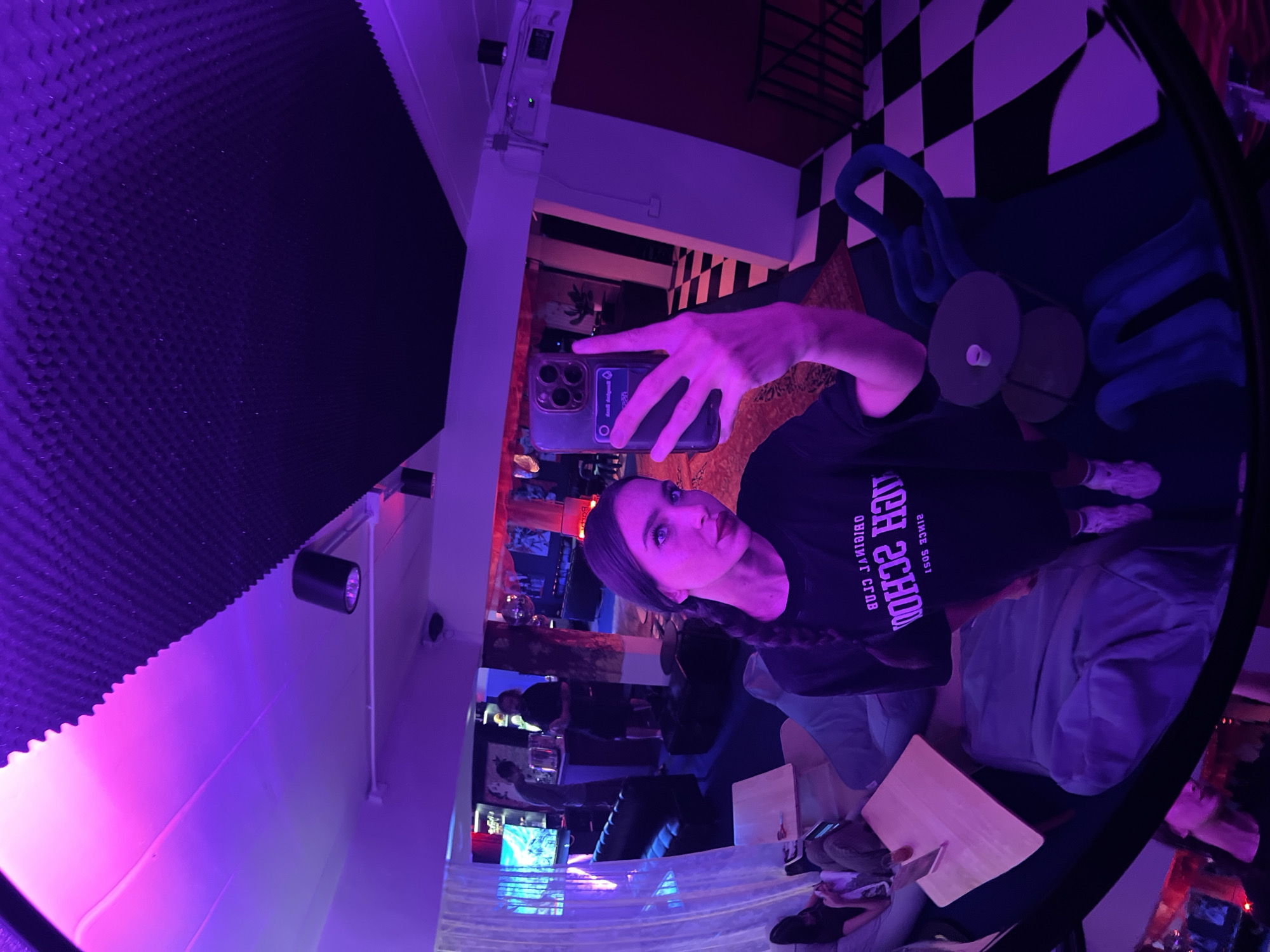 A selfie reflection in a round mirror taken at a venue in Phuket City Municipality, Thailand. The woman has her hair in a braid and is wearing a t-shirt with text. She holds a phone up to take the picture. The room contains a black and white checkered floor, a blue octopus sculpture, sound dampening material on the wall, and a bar area with seating. The lighting is a deep purple tone.