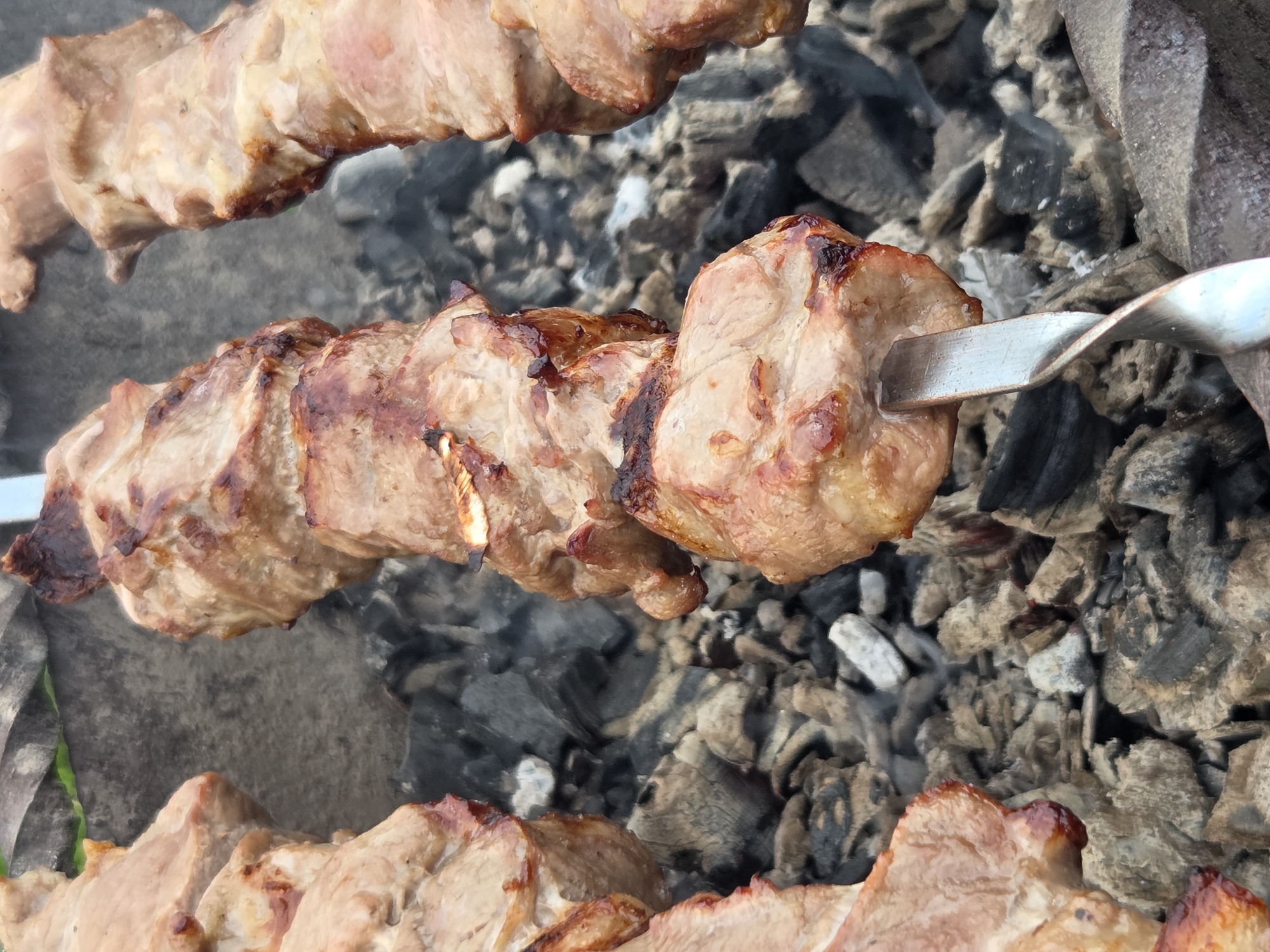 This image shows three skewers of meat cooking over hot coals. The meat is browned and appears to be well-cooked. The skewers are made of metal and are positioned over a bed of gray and black coals. The background is blurred, but it appears to be an outdoor setting. There are no people visible in the image. The location in the city of Frolovo, Russia, is not evident from the image itself.
