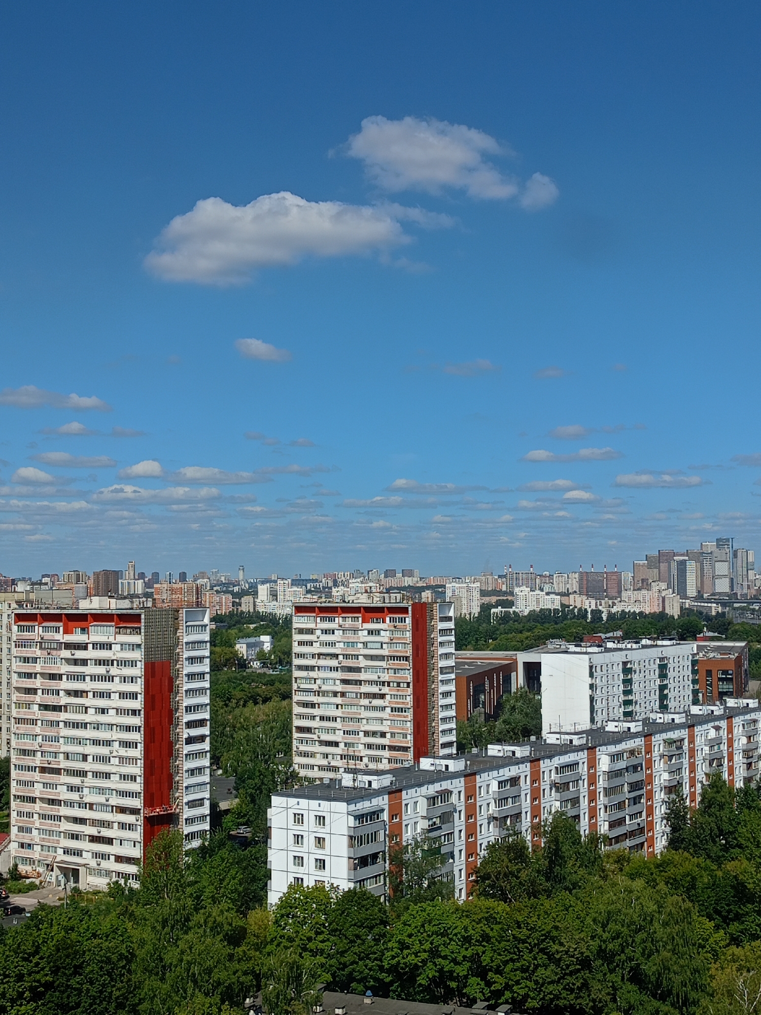 The image captures a panoramic view of a city skyline on a clear day. The foreground is dominated by lush green trees, obscuring the lower portions of the buildings. In the mid-ground, a series of multi-story apartment buildings are visible. Some of these buildings are tall and slender with white and red facades, featuring numerous windows and balconies. Other buildings are longer and lower, also with a pattern of windows and balconies, and some have orange accents. In the background, the city extends further with a dense collection of buildings of varying heights and architectural styles, some appearing to be modern skyscrapers. The sky above is a vibrant blue, dotted with scattered white clouds. The overall impression is of a densely populated urban environment with a mix of residential and possibly commercial structures, set against a bright, sunny sky. There are no visible people or animals in the frame.