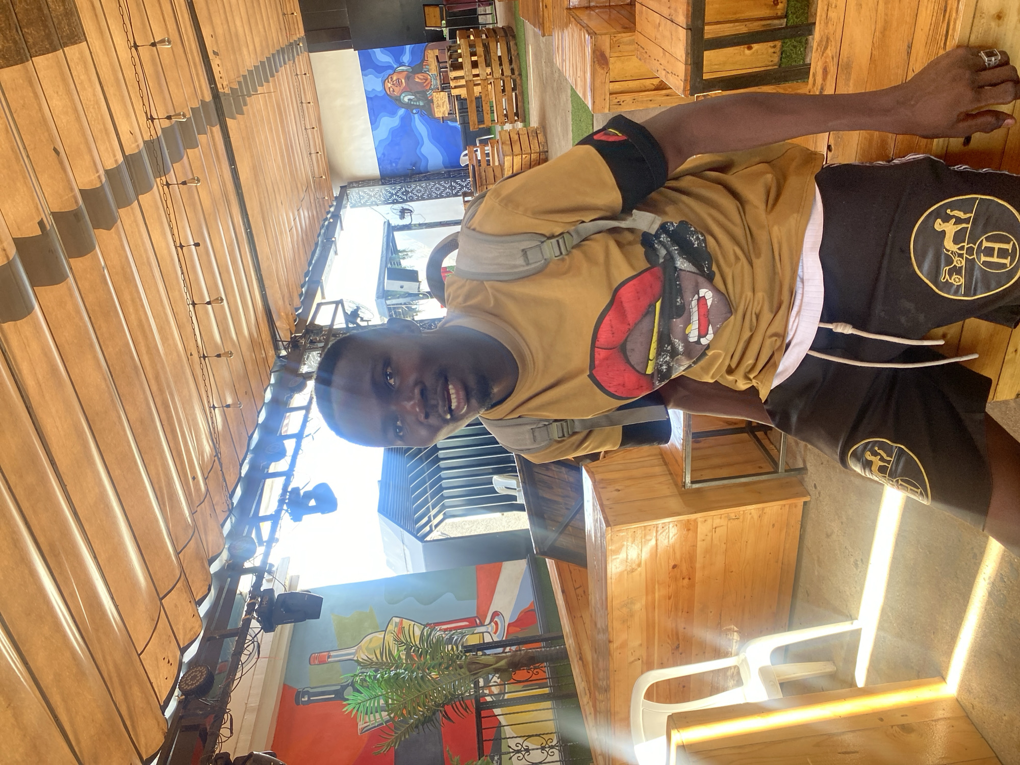 The image depicts a male individual within an outdoor or semi-outdoor establishment, identified as being in Howayi, Nigeria. The subject, positioned centrally, is a dark-skinned male looking directly at the camera with a slight smile, mouth slightly ajar. He wears a mustard yellow short-sleeved t-shirt featuring a cartoonish graphic of a face with prominent red lips and sunglasses. A grey backpack is slung over his shoulders. He is wearing black shorts with a gold circular emblem depicting a horse-drawn carriage and the letter "H" on each thigh, and a white undershirt is visible beneath his t-shirt. A black armband with a small yellow/red design is visible on his right arm, and he wears a silver-colored ring on the pinky finger of his left hand. He appears to be seated on a wooden bench.
The environment features a light brown corrugated fabric canopy overhead, supported by metal beams from which stage lights and decorative Edison-style light bulbs hang. Strong sunlight enters from the left and front, creating bright lens flares and distinct shadows on the concrete floor. The walls are adorned with murals: the left wall displays bottles, a glass, and a palm plant, while the right wall features a blue-toned mural of a person with headphones. The seating arrangements consist primarily of light-colored wooden structures resembling benches and tables, constructed from stacked or joined planks. A single white plastic chair is visible on the left. Speakers are mounted on the overhead structure, and a dark structure with vertical slats is visible in the background.