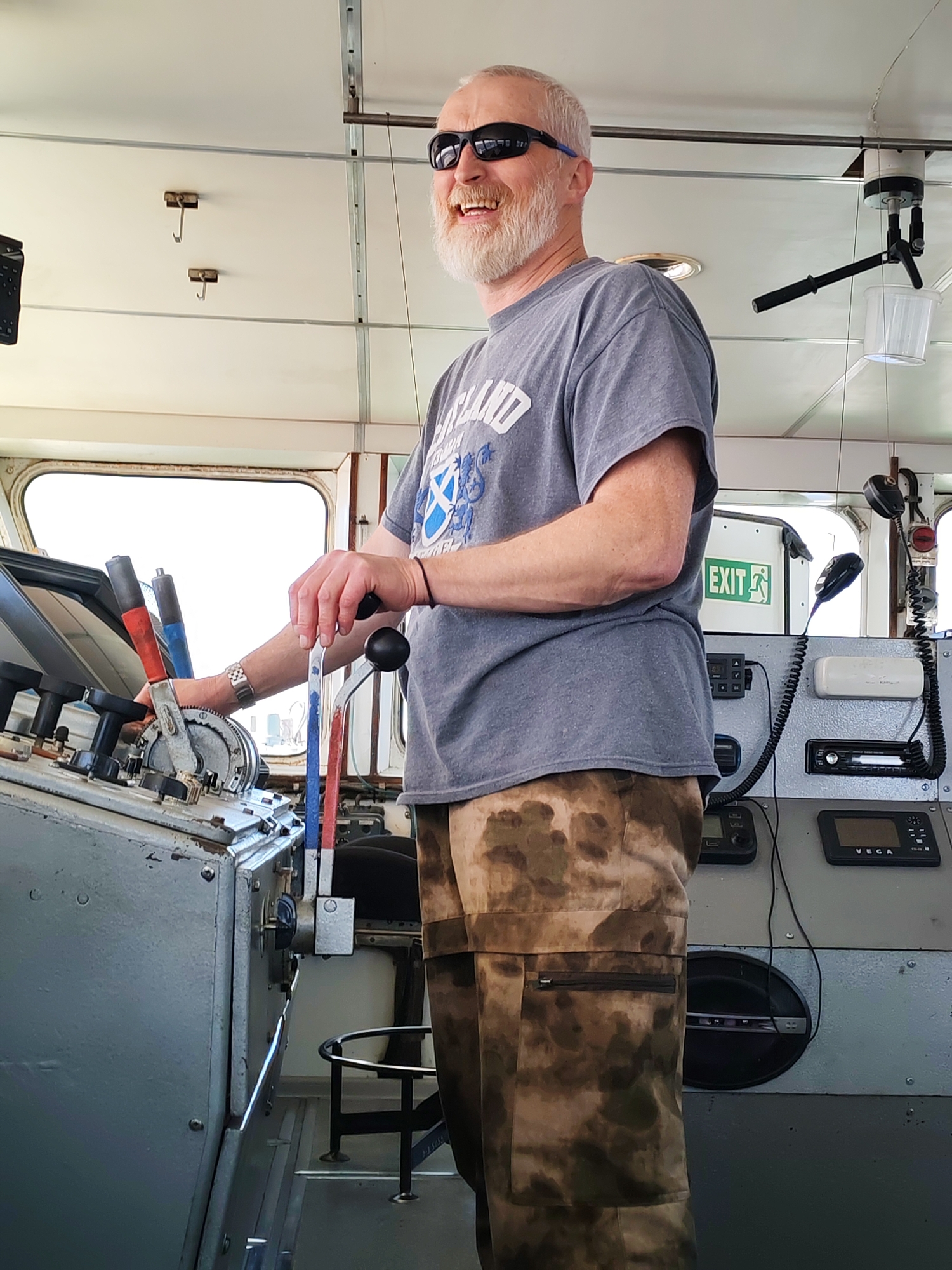 A middle-aged man with a white beard and sunglasses stands at the controls of a vessel. He is wearing a gray t-shirt and camouflage pants. He has his hands on the ship's controls. Behind him are communication devices, a microphone, and an exit sign. The setting appears to be the interior of a ship or boat. There is no evidence in the image to suggest the location is Koprino, Russia.
