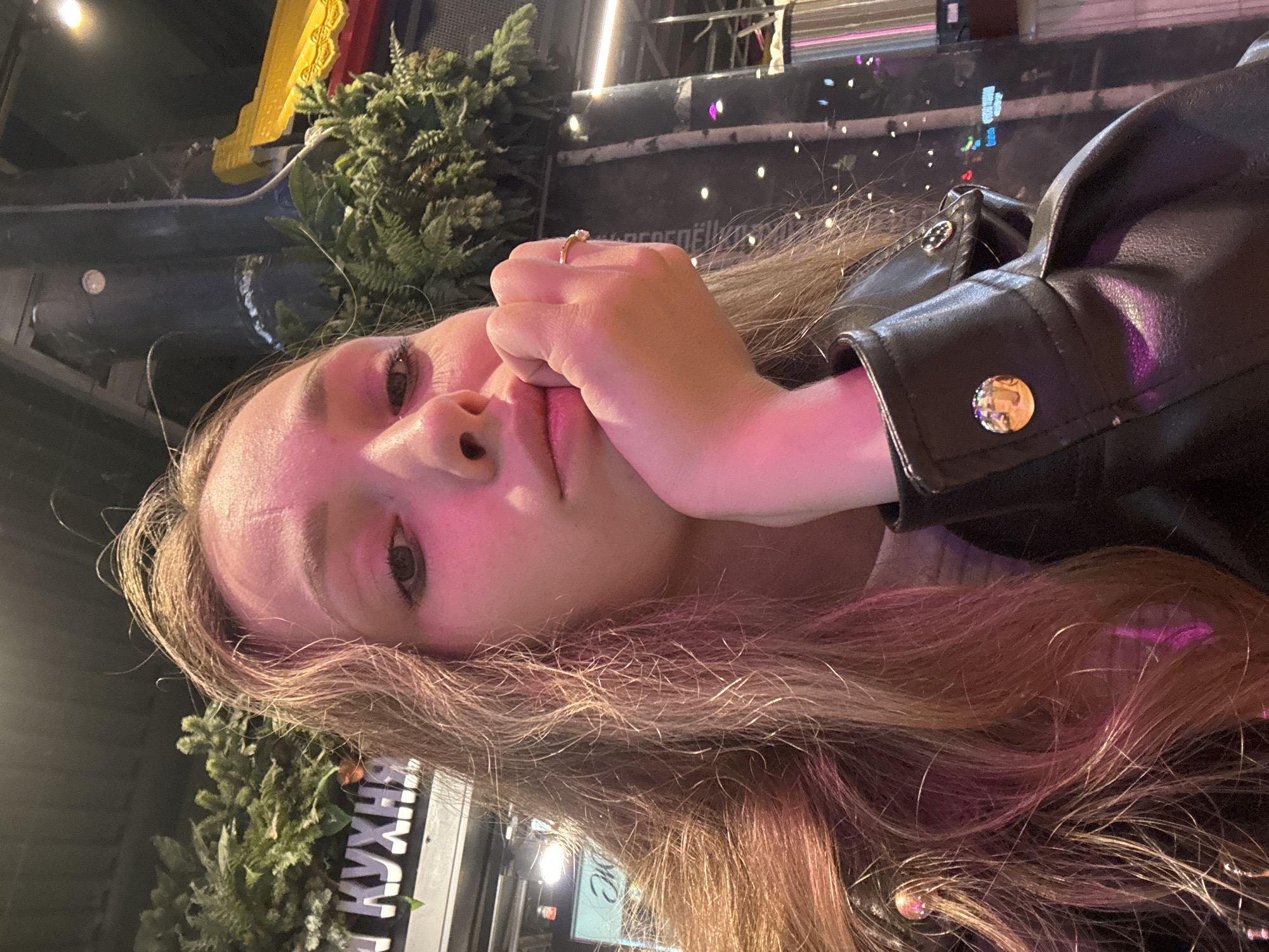 A medium, close-up, portrait-style photograph shows a woman with light skin and long, light-brown hair who is wearing a black leather jacket. She has a gold ring on her finger, and is holding her hand to her face. The lighting is soft and pinkish. Behind her are indistinct blurred objects, including some greenery that could be artificial. A sign with Cyrillic writing appears in the background. The general appearance of the scene suggests it is in Yekaterinburg, Russia.