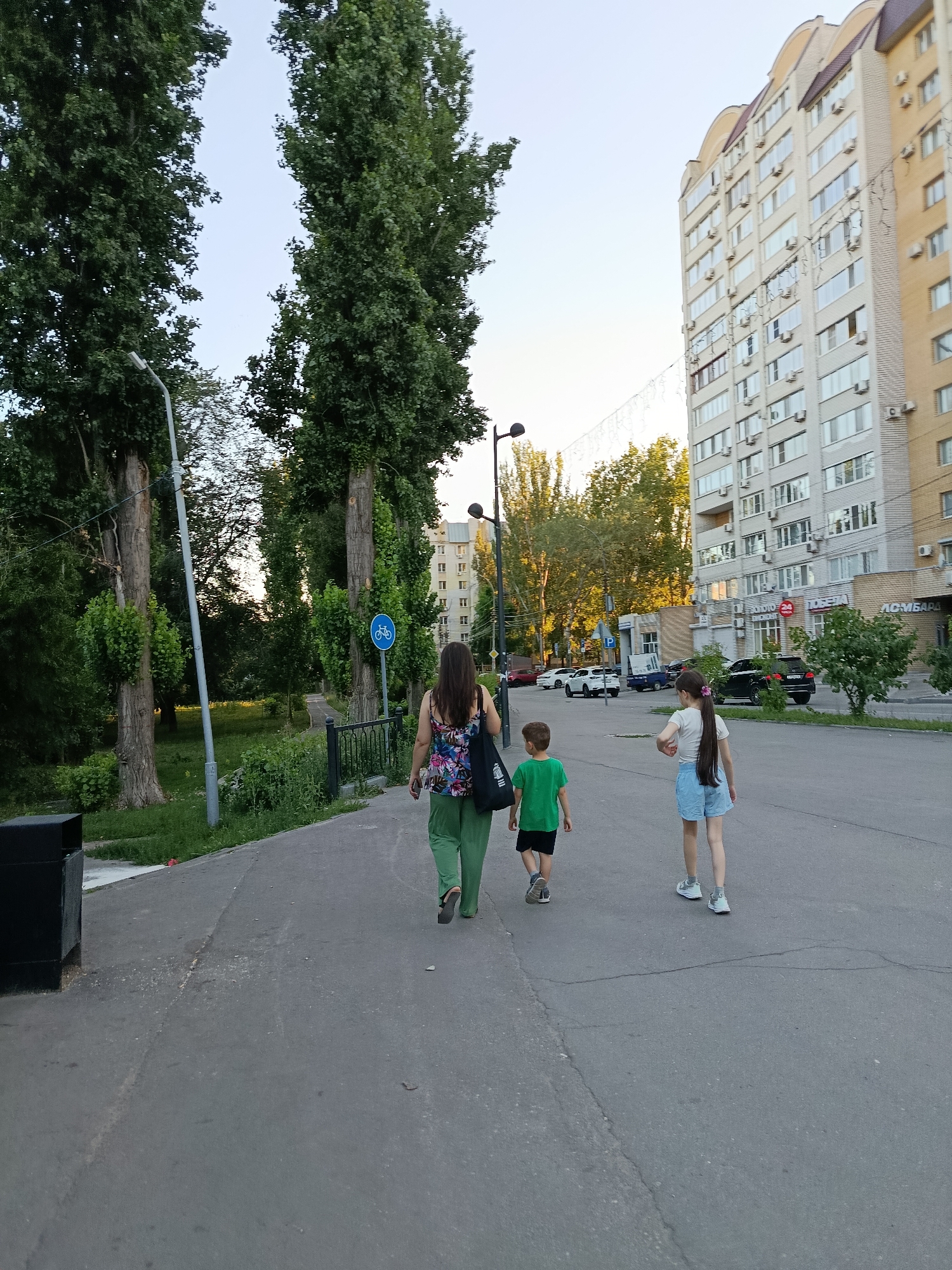 A woman and two children, a boy and a girl, are walking away from the camera on a paved path next to a street. The woman is wearing green pants and a colorful floral top. The boy is wearing a green t-shirt and black shorts. The girl is wearing a white t-shirt and light blue shorts. They are walking in a suburban area with tall trees and a multi-story apartment building in the background. The sun is low in the sky, casting long shadows, indicating it is late afternoon or early evening. The weather appears to be pleasant and warm. There is a bicycle lane sign visible next to the path. Visible text includes "ЛОМБАРД" and "ПОБЕДА 24" on the apartment building. The overall mood seems relaxed and casual, suggesting a family outing or a stroll.