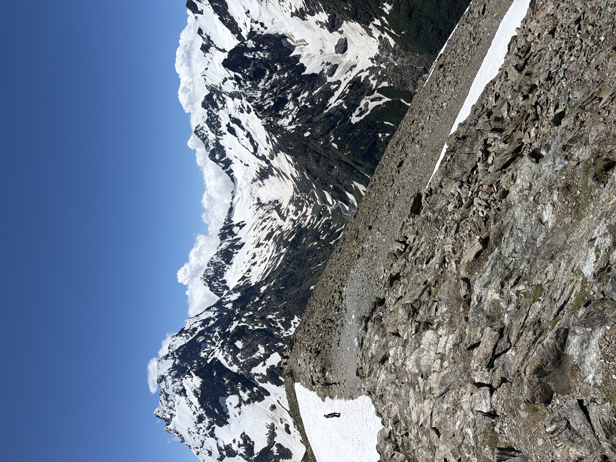 The image depicts a mountainous landscape with snow-capped peaks under a clear blue sky. The foreground features a rocky, barren ridge, with patches of snow scattered on its surface. Two individuals are visible on the ridge; one is standing on a patch of snow further up the ridge, and another appears to be walking in the distance. The midground showcases a vast valley with steep, snow-covered slopes and dark, rocky outcrops. The background is dominated by a range of jagged, snow-covered mountains under a bright blue sky with scattered white clouds. The scene suggests a remote, high-altitude environment suitable for hiking or mountaineering. The general location is inferred to be within the Верхне-Тебердинское сельское поселение, Russia, a region known for its mountainous terrain and natural beauty.