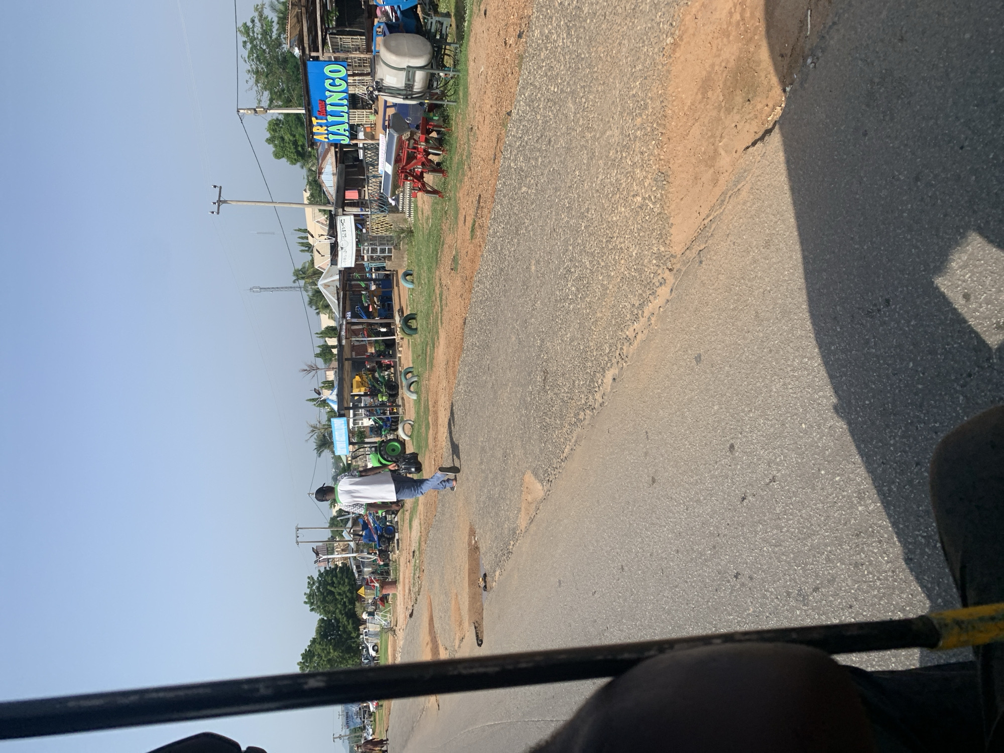 The image depicts a roadside scene under a clear blue sky. In the foreground, a black pole, partially visible, suggests the perspective is from a vehicle. A person walks away from the camera on a cracked asphalt road. They are wearing a white shirt with a green design and blue jeans. To the right of the road, there is a sandy shoulder with some scattered debris and a shadow cast by an unseen object. Beyond the shoulder, various structures are visible, including small shops with corrugated metal roofs. Agricultural equipment, such as tractors and what appear to be seed drills, are displayed in front of these establishments. A prominent blue sign with yellow lettering reads "ART HOME JALINGO". In the background, trees and utility poles are scattered across the landscape.