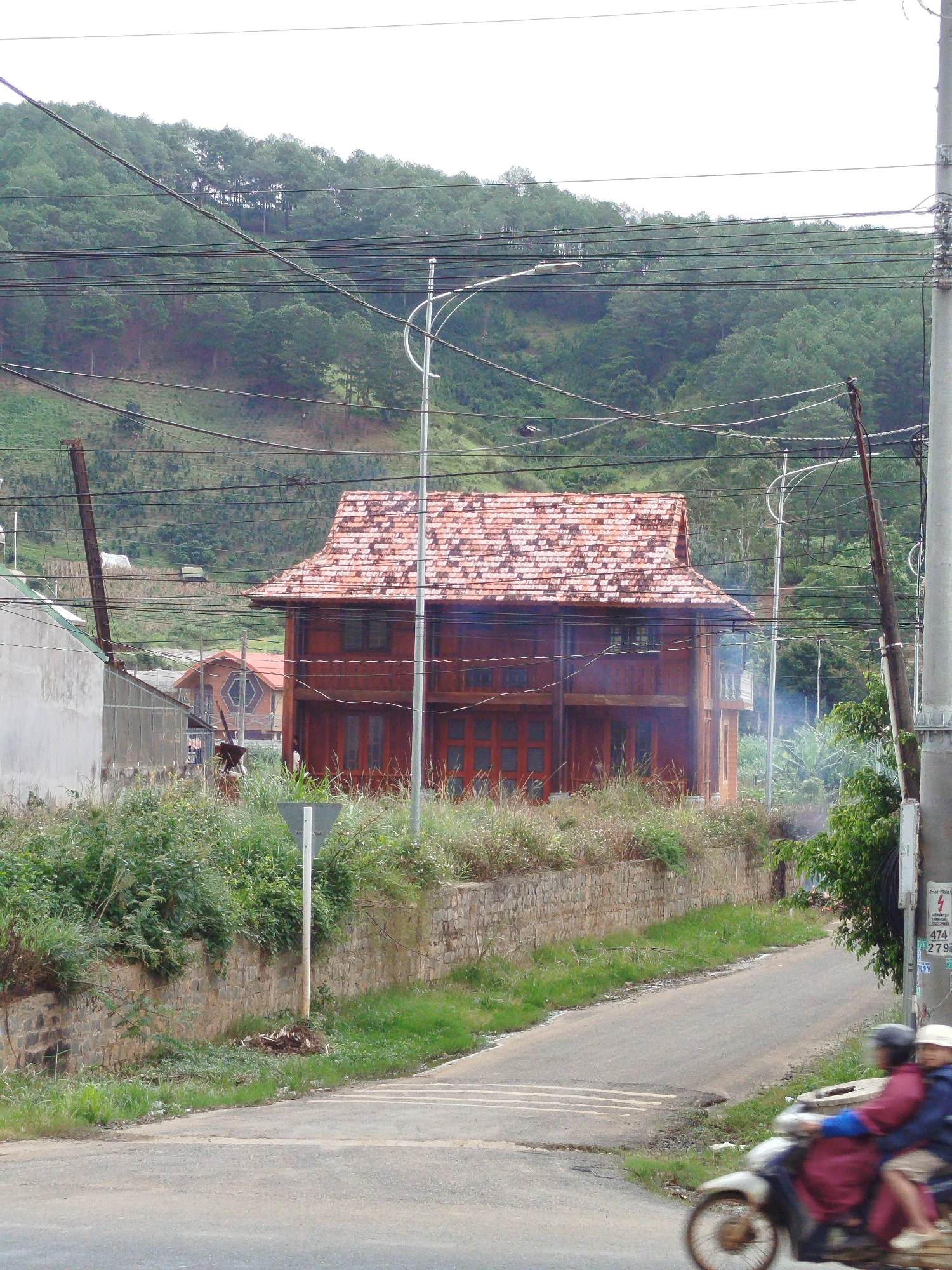 The image depicts a street scene in K'Long, Vietnam, characterized by a prominent wooden house with a red tile roof situated amidst lush vegetation and a hillside covered in pine trees. Power lines crisscross the sky, supported by wooden and concrete poles. To the left of the wooden house, a lighter-colored building with a sloped roof is partially visible, and further in the background, another house with a distinct hexagonal window is seen. A rough stone wall runs along the side of the road, overgrown with grass and plants. The road itself is paved and appears to be a two-lane street. In the foreground, two individuals are riding a motorcycle, their forms blurred due to motion. The overall atmosphere is that of a semi-rural or suburban area, with a blend of residential structures and natural elements.