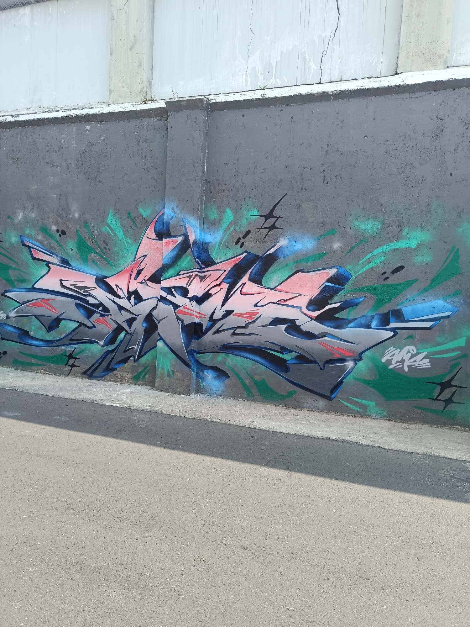 The image shows a graffiti mural on a concrete wall. The mural features abstract, stylized lettering in shades of pink, gray, and blue, with green spray-painted accents and black splatters. The wall is gray and appears to be part of an outdoor structure, possibly a bridge underpass or a retaining wall. In the foreground, there is an asphalt surface, suggesting a road or walkway. The background shows the upper portion of a light-colored concrete wall with some cracking. The scene appears to be in an urban environment in Yogyakarta, Indonesia, given the style of construction and the graffiti. There are no people or animals visible in the image.