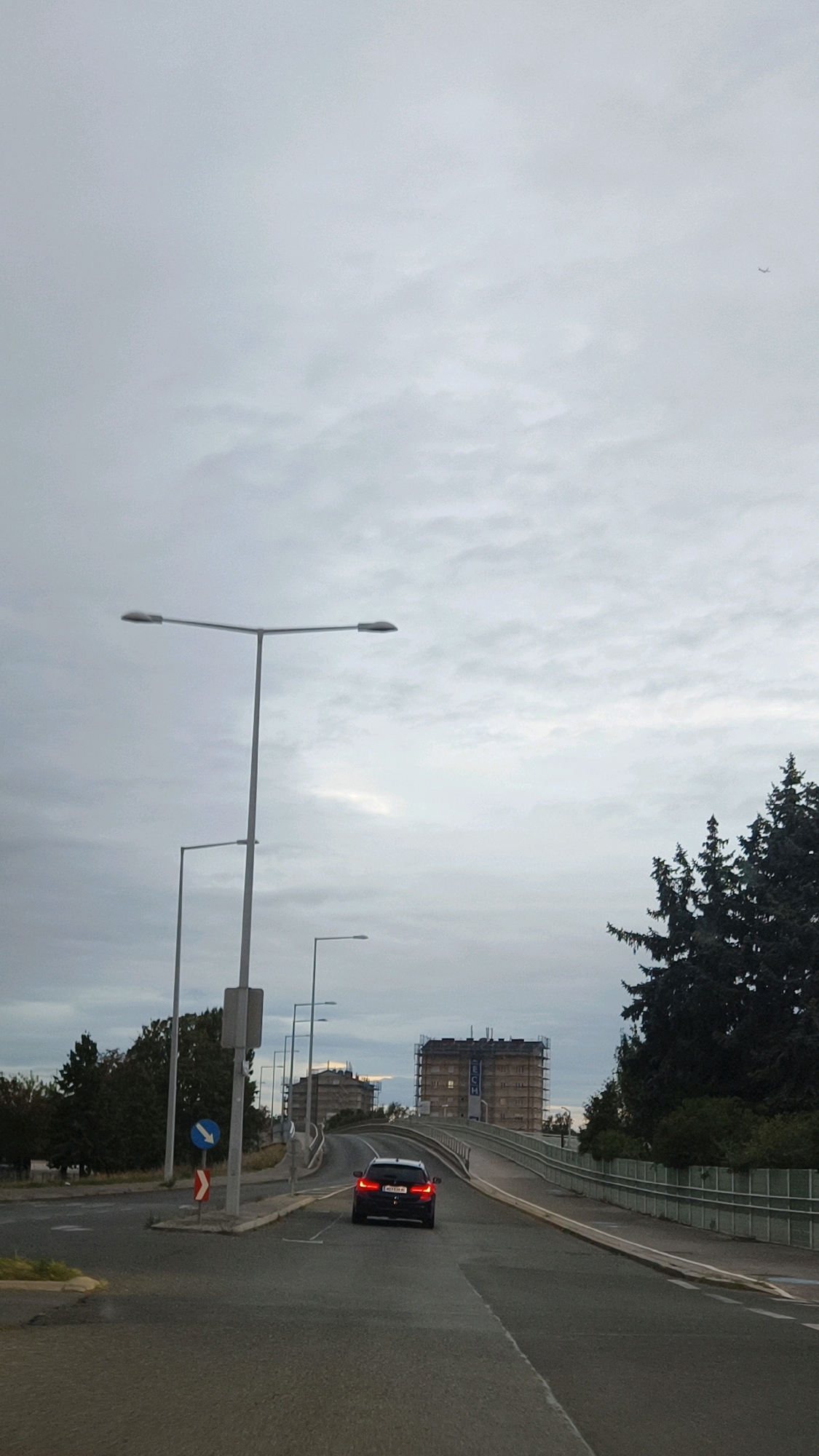 A dark sedan drives away from the camera on a slightly overcast day in Vienna, Austria. The car is on a road that curves upwards towards a bridge. On the right side of the road are tall evergreen trees. In the background, two large buildings are under construction. The sky is cloudy and a small airplane is visible in the upper right corner of the image.