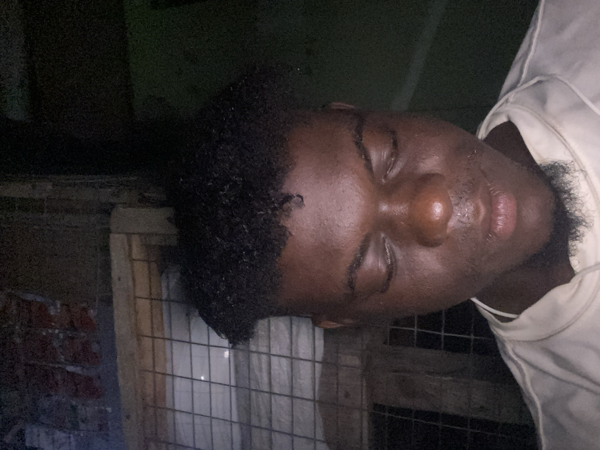 A dark-skinned man with a beard and curly hair is sleeping inside a wooden cage. The location is Unknown, Unknown. The cage is located in a dark area, possibly a basement or storage room. The man is wearing a white t-shirt. There is a brick wall near the cage.