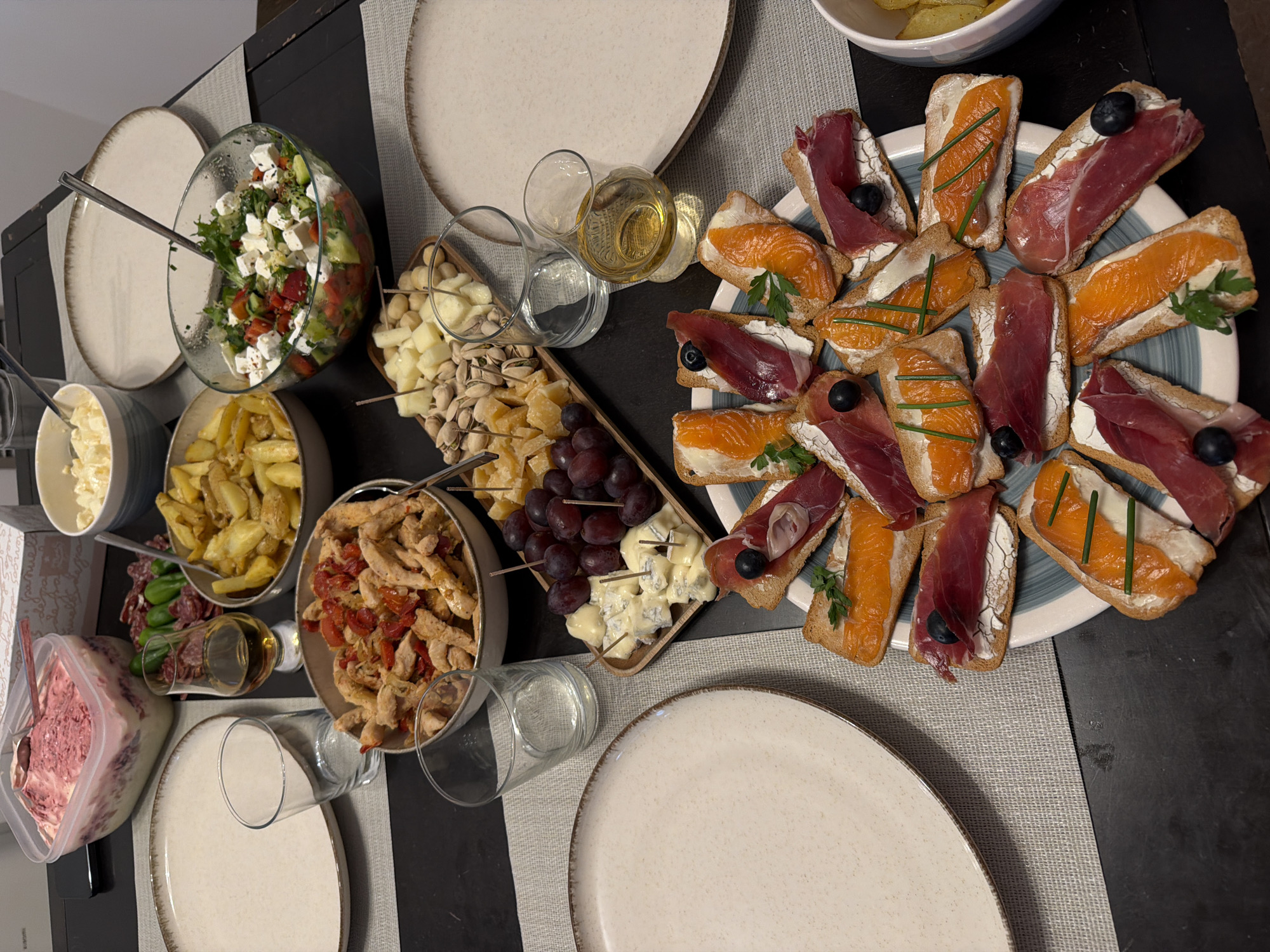 A high-angle shot depicts a table laden with food. Plates are set around the table, which is covered with a striped gray placemat. The food includes a salad in a glass bowl, a bowl of potatoes, a bowl of chicken and tomatoes, and a container of what appears to be beetroot salad. A wooden platter holds cheese, grapes, nuts, and skewers of cheese. A large plate is covered in bruschetta topped with salmon and prosciutto. Glasses and a wine bottle are also visible. The location cannot be determined with the provided visual information. There is no indication it is in Valencia, Spain. There are no people visible in the image.