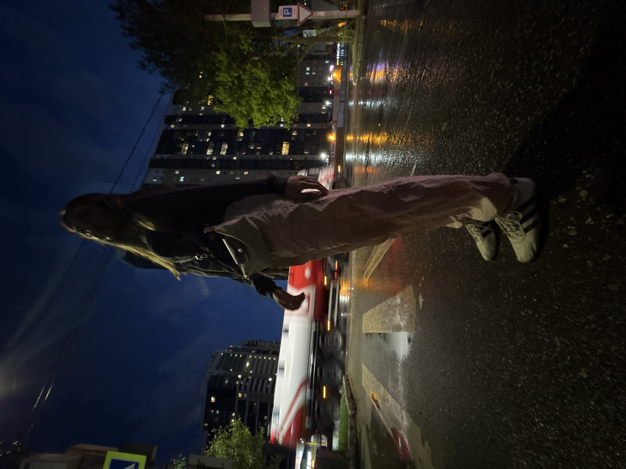 A nighttime scene in Perm, Russia, shows a person standing near a wet street with light reflections. The person is wearing light-colored pants, a dark jacket, and white Adidas sneakers. A large building with many illuminated windows is visible in the background. A red and white vehicle appears to be moving quickly in the street. A parking sign is also visible in the background.